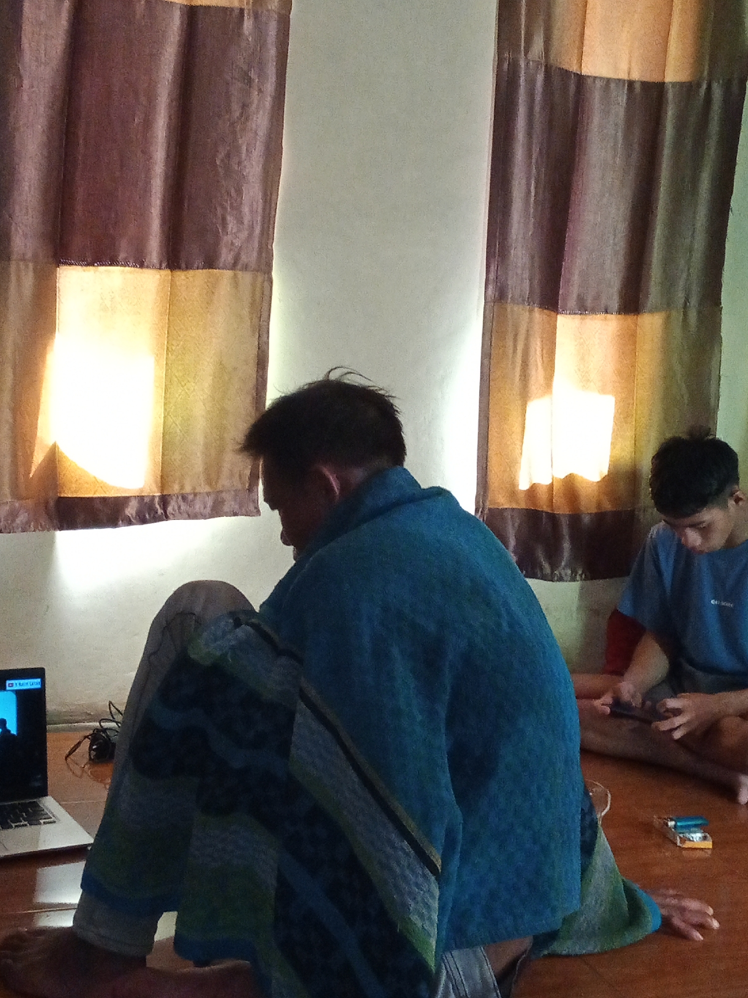 The image depicts an indoor scene, possibly a living room in a residence located in Subang, Indonesia. Two individuals are present. The person in the foreground, facing away from the camera, is wearing a blue blanket or robe. This individual is seated on the floor, with a laptop to their left and what appears to be a charging cable. In the background, a second person, a younger male, is also seated on the floor and is engaged with a mobile phone. He is wearing a blue t-shirt with red sleeves. The environment includes two large windows covered by curtains that have a striped pattern of dark brown and lighter tan/yellow hues. Sunlight is filtering through these curtains, casting bright patches of light onto the wall and floor. On the floor near the second individual, there is a small object, possibly a cigarette pack with a lighter on top. The floor appears to be made of polished wood.