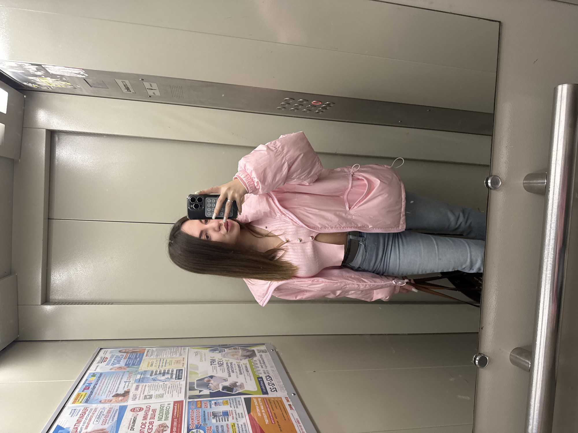 A young woman stands in an elevator, reflected in a mirror, taking a selfie with her phone. She is wearing a pink jacket and jeans. The elevator interior is a light gray color. There is a metal control panel above her head and a newspaper advertisement panel on the wall below her. The advertisement contains phone numbers including 8 (800) 623-3333 and mentions "Всолярка" and "OTEHNW BOUC." There are other Cyrillic words and phrases on the advertisement.