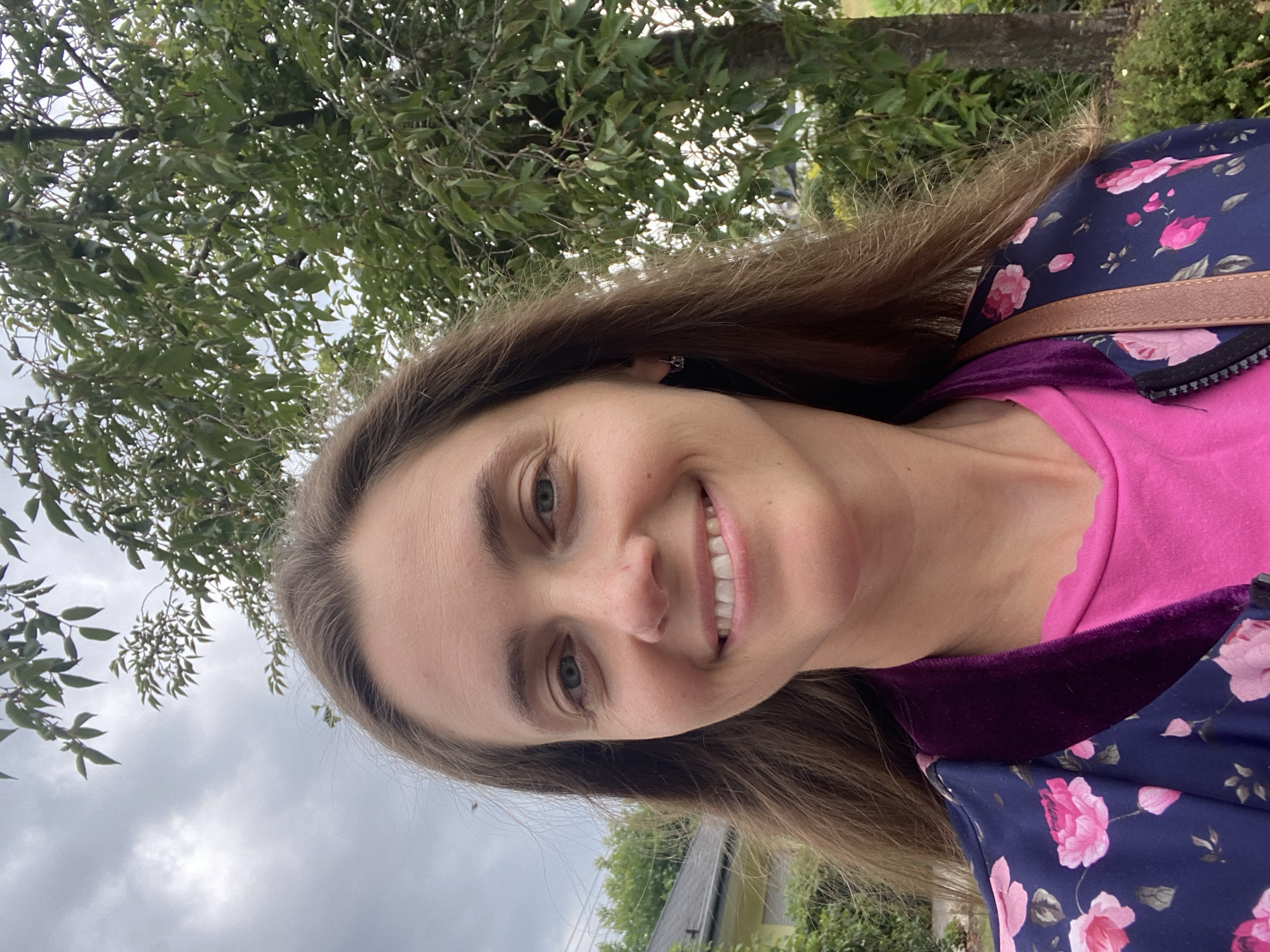 A woman is captured in a selfie outdoors with a cloudy sky and trees in the background. She is smiling directly at the camera, her long brown hair cascading down her shoulders. She is wearing a vibrant pink t-shirt and a navy blue jacket adorned with a floral pattern of pink roses. The jacket also features a dark purple velvet collar. To the left of the woman, a portion of a house with a dark roof and yellow siding is visible, partially obscured by greenery. The scene appears to be set in a residential area of Flammersfeld, Germany.