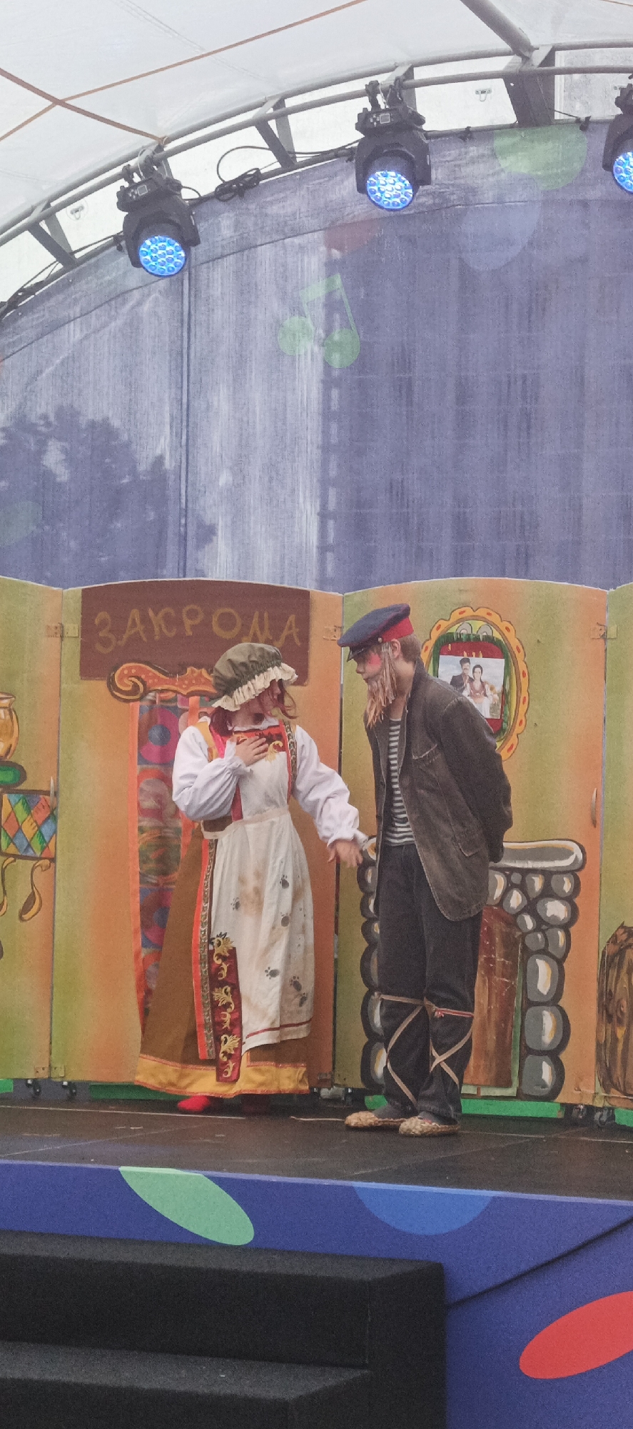 The image shows two people in costume on a stage, likely performing a play or theatrical production. The backdrop depicts a rustic, colorful setting, with painted wooden panels that suggest a stylized village or interior scene.
The person on the left is dressed in traditional folk attire. They wear a white blouse with puffy sleeves, a long, ornate skirt or dress in brown and red with embroidered trim, and a white apron. A ruffled, green head covering frames their face. Their right hand is held to their chest, and their gaze is directed downwards, conveying a sense of tenderness or perhaps sorrow.
The person on the right is dressed as a more rugged character. They wear a striped shirt, a dark jacket, dark trousers, and a cap. Their face has a painted beard or stubble and rosy cheeks. Their legs are wrapped with crisscrossing straps, and they wear simple, straw-like footwear. They are holding the left person's hand, and their head is slightly bowed, looking towards the other performer.
The stage has a painted fireplace on the right side and a sign above the left person that reads "ЗАКРОМА" (ZAKROMA), which translates to "granaries" or "storehouses." A framed picture of a couple is visible on the backdrop behind the right performer.
Above the stage, stage lighting is visible, casting blue light. The overall scene appears to be an outdoor event given the lighting structure and the open-air feel of the backdrop. The mood seems to be dramatic or heartfelt, suggested by the performers' poses and expressions. The location context is Izhevsk, Russia.