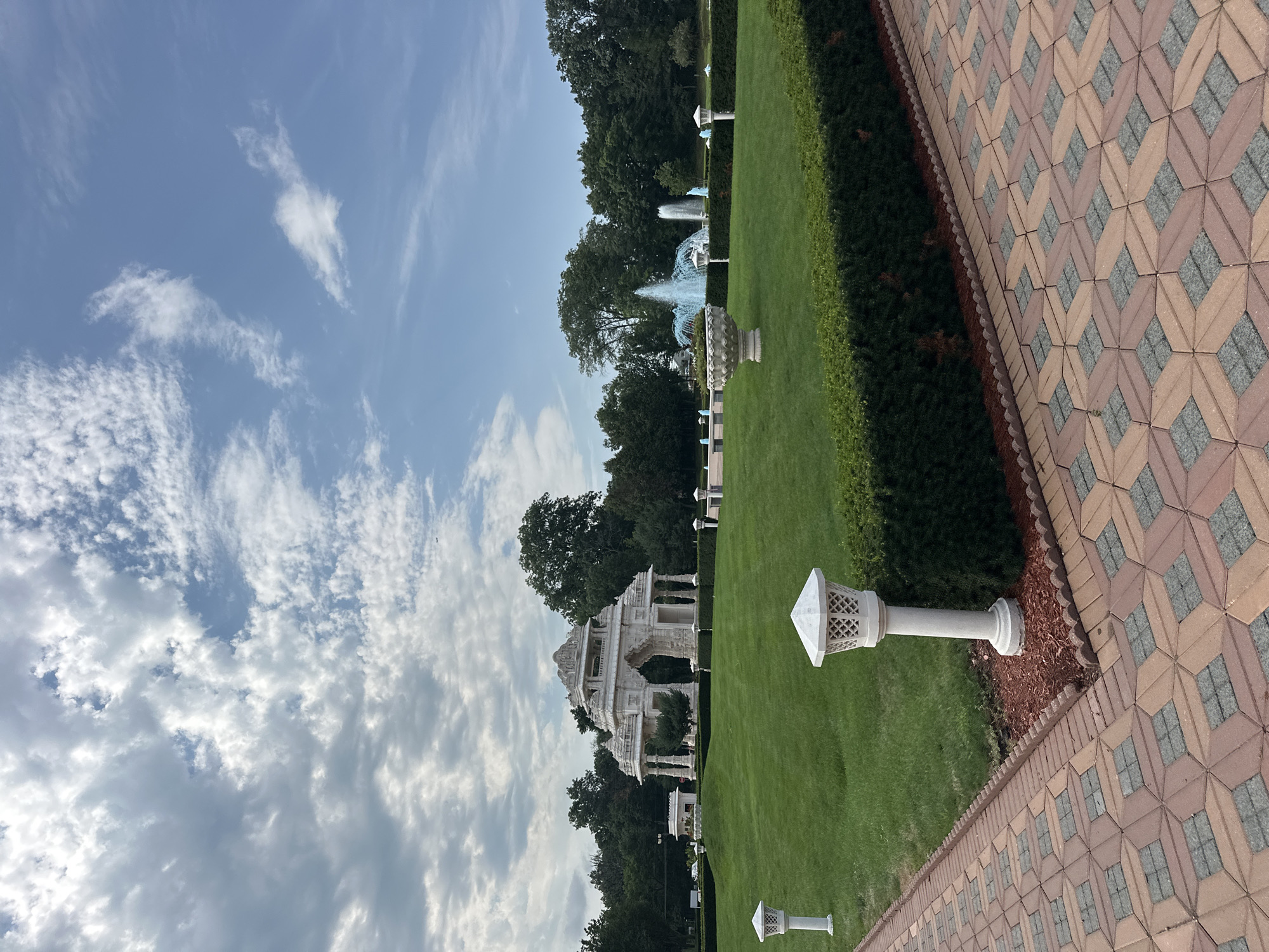 The image depicts the exterior of a Hindu temple located in Bartlett, Illinois, United States of America. In the foreground, a patterned brick walkway leads towards a manicured lawn bordered by a neatly trimmed hedge. Several ornate white lampposts are strategically placed along the lawn's edge.
Dominating the mid-ground is a large, white, intricately carved structure resembling an archway or gateway, characteristic of traditional Hindu architecture. Behind this structure, a series of fountains spray bright blue water into the air. The background is filled with lush green trees, suggesting a park-like setting. The sky above is a mix of blue sky and scattered white clouds. The overall scene conveys a sense of grandeur and tranquility associated with a religious site and its surrounding gardens.