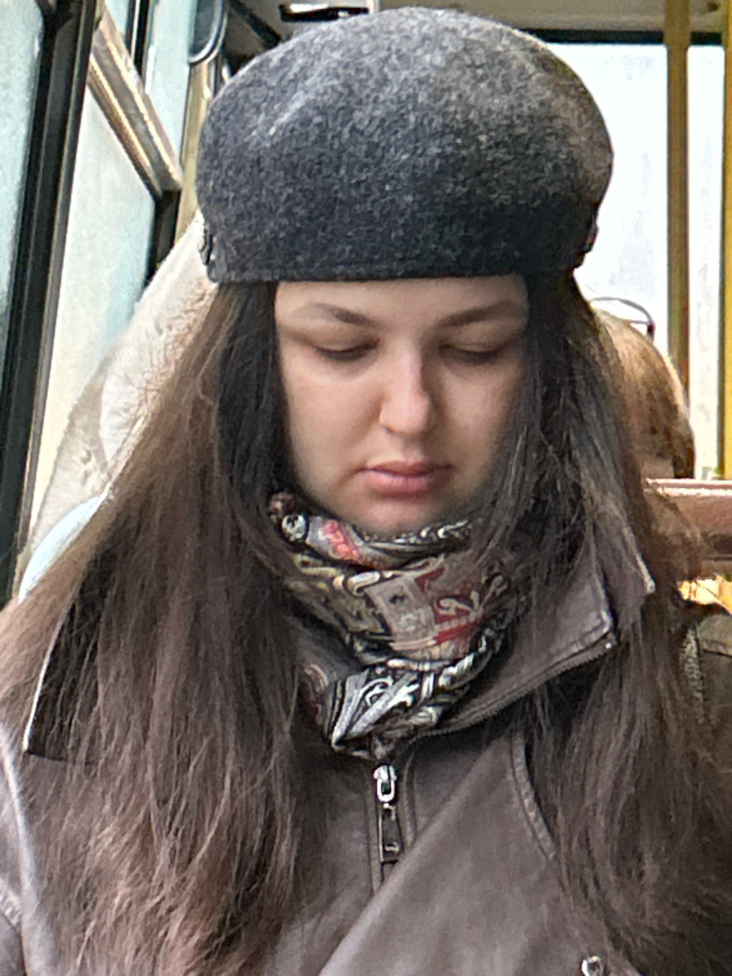 A woman wearing a dark grey beret and a patterned scarf is visible from the chest up. She has long, dark brown hair that falls over her shoulders and down her back. She is wearing a brown leather jacket. The background appears to be the interior of a public transport vehicle, possibly a bus or tram, with windows and metal framing. The lighting is diffused, suggesting an indoor setting. The image is a medium shot, focusing on the woman's face and upper body. It is not possible to determine the specific location within the city of Tomsk, Russia, based on the visual information provided.
