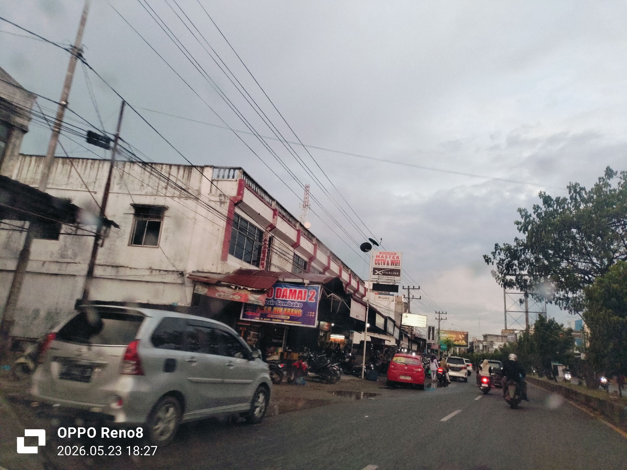 The image shows a street scene in Baro, Indonesia. The view is taken from a moving vehicle, likely a car or motorcycle. In the foreground, there is a silver minivan and a portion of the road. Further along the road, there are several motorcycles and cars moving in both directions, suggesting moderate traffic. On the left side of the road, there is a two-story commercial building with several storefronts. One storefront displays a large sign that says "DAMAI 2" which is a grocery store selling basic goods. Another sign advertises "MASTER CCTV & WIFI STARLINK." There are pedestrians on the sidewalks. The weather appears overcast, with a cloudy sky. Power lines and utility poles are visible overhead. The image also includes the camera information at the bottom left corner, showing "OPPO Reno8" and the date "2026.05.23 18:27". The scene suggests a typical urban commercial area.
