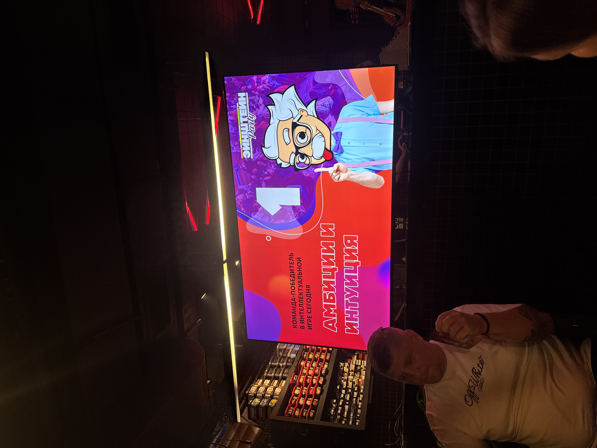 The image captures a man wearing a white "Off-White" t-shirt in a dark setting, likely indoors. In the background, a large vertical screen displays an advertisement or announcement in Russian. The screen features a cartoon image of a character resembling a scientist, with text that reads "ЭИНШТЕЙН" (Einstein) and "КОМАНДА-ПОБЕДИТЕЛЬ В ИНТЕЛЛЕКТУАЛЬНОЙ ИГРЕ СЕГОДНЯ" (Winning team in today's intellectual game). Underneath, the words "АМБИЦИИ И ИНТУИЦИЯ" (Ambition and Intuition) are written. There are also trays containing boxes that appear to be playing cards. The location of this image is likely Kuta Selatan, Indonesia, based on the instruction.