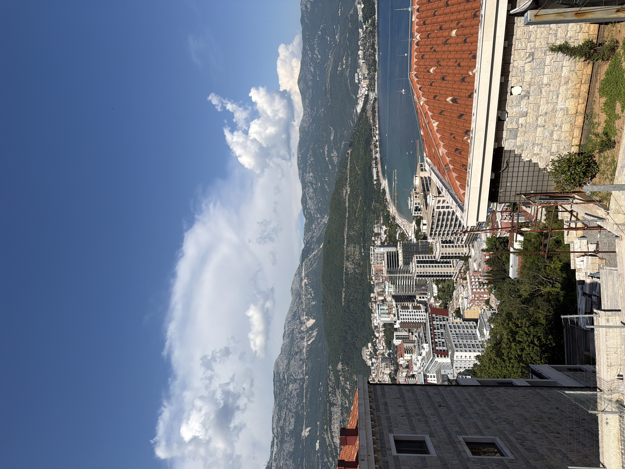 The image is a high-angle, wide shot of the coastal city of Budva, Montenegro. In the foreground, on the right, is a building with a red tiled roof. The building is clad in stone blocks, with a visible section of mosaic tile work. To the left of this building, a stone-clad wall with two windows is visible. Further down the slope, construction materials are piled up.
In the mid-ground, a densely packed city skyline is visible, comprised of numerous multi-story buildings, many with white facades. Some buildings are modern, with glass elements, while others appear older. Green trees are interspersed among the buildings.
The background features a bay with calm, dark blue water. A beach lined with umbrellas and sunbathers is visible along the coastline. Several small boats are anchored in the bay. Beyond the city and the bay, a range of rugged, green mountains rises, with some lighter-colored rock faces visible. The sky above is a vibrant blue, with scattered white clouds, some of which are large and puffy, casting shadows on the mountains. The overall impression is a sunny day in a coastal city.