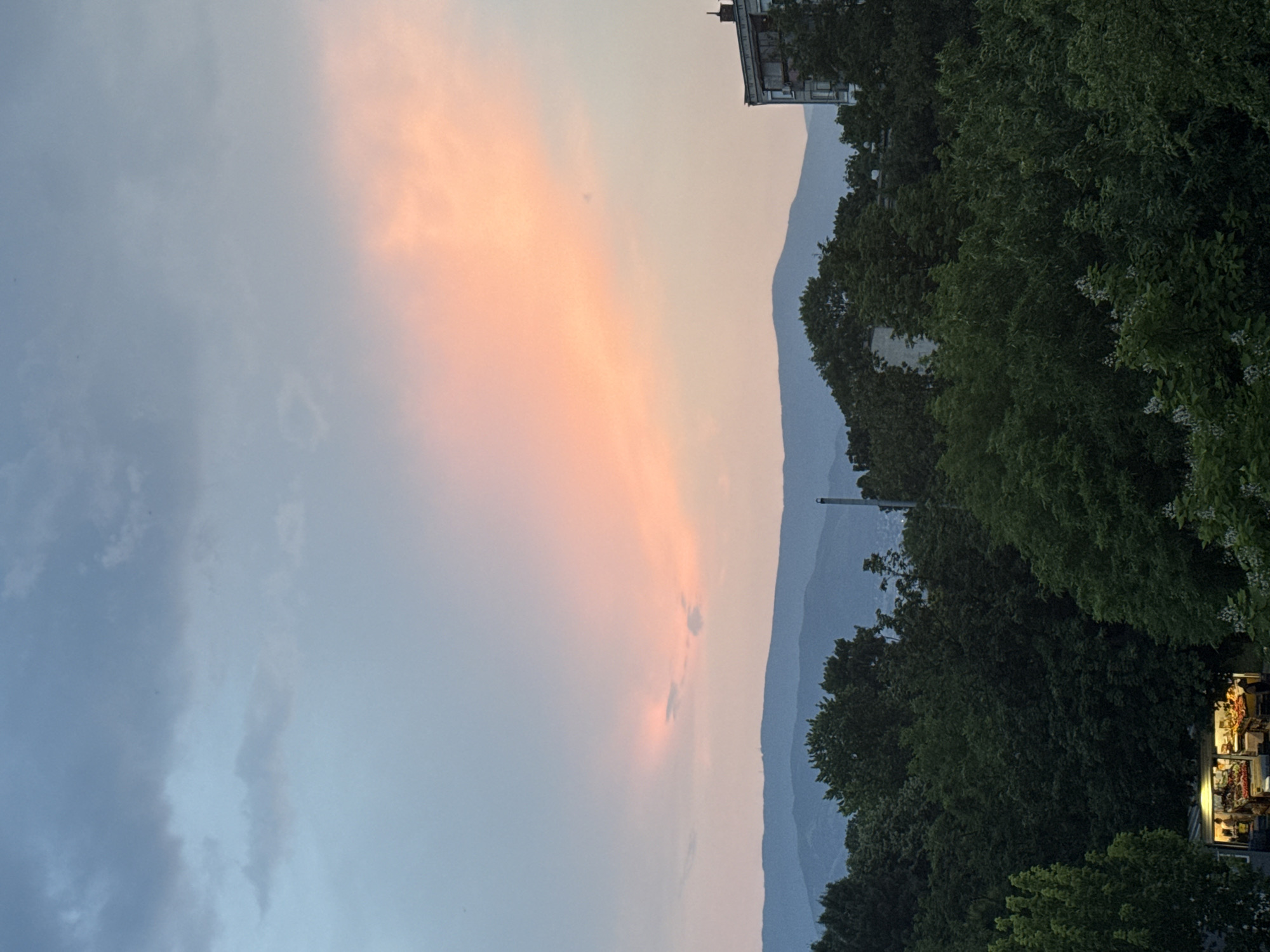 The image depicts a twilight scene over a landscape featuring natural and urban elements, consistent with a view from Plovdiv, Bulgaria, with the Rhodope Mountains in the distance. The upper portion of the frame is dominated by a sky transitioning from a pale blue-grey at the top, with scattered dark grey stratocumulus clouds, to a prominent horizontal band of soft orange and pink hues across the mid-sky, indicative of sunset.
A continuous, undulating range of dark blue-grey mountains forms the distant horizon, silhouetted against the lighter sky. Below the mountains, a dense canopy of dark green deciduous trees occupies the lower half of the image, obscuring most ground-level details.
In the lower left quadrant, partially obscured by trees, a brightly illuminated, single-story market stall or kiosk is visible. Warm artificial light emanates from within, highlighting various items, consistent with produce (fruits and vegetables), arranged on shelves or tables. Two indistinct human figures are discernible inside the illuminated stall.
In the upper right quadrant, the top three stories and roofline of a grey, multi-story residential building are visible above the tree line. The building has a flat roof with minor architectural detailing and a closed balcony structure. Slightly to the left of this building, a tall, slender, dark grey cylindrical structure, resembling an industrial chimney or vent pipe, rises vertically above the tree canopy.
The overall lighting suggests dusk, with natural light fading and artificial light sources becoming prominent in the foreground. No specific actions or interactions beyond the presence of people in the market stall are discernible.