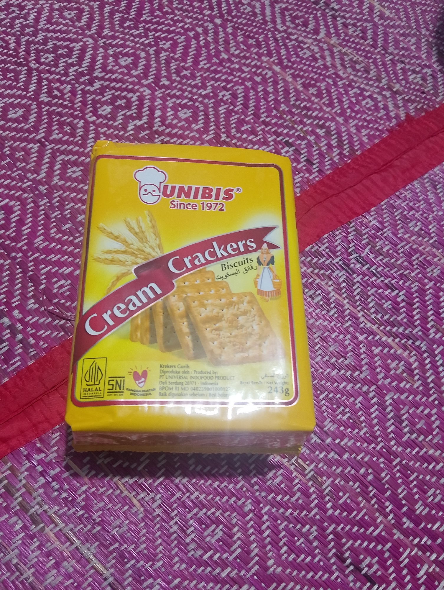 A yellow package of Unibis Cream Crackers is lying on a woven mat. The mat is made of pink and white fibers woven in a diagonal pattern. A red ribbon is present on the right side of the mat, running diagonally from the top right to the bottom left. The package is positioned horizontally, with its front facing the viewer. The branding "UNIBIS Since 1972" is prominently displayed in white text on a red banner. Below this, "Cream Crackers" is written in white text. Arabic text is also visible on the package. There are images of wheat stalks and a woman in traditional clothing carrying a basket of biscuits. Halal and SNI (Indonesian National Standard) logos are present at the bottom left of the package, along with text indicating the product is made in Deli Serdang 20371, Indonesia. The net weight is listed as 243g.