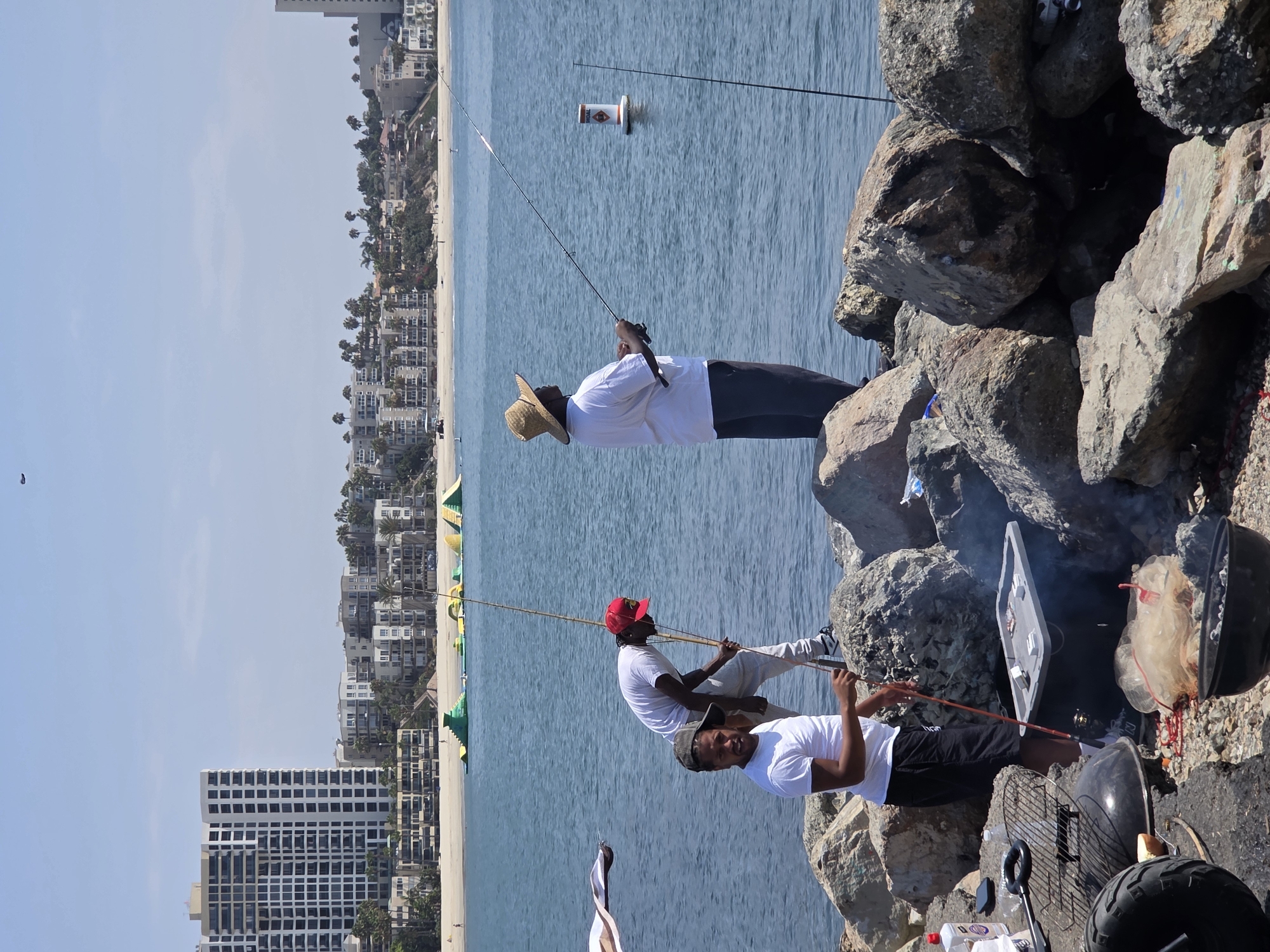 Three men are fishing on a rocky breakwater in the city of Long Beach, California. In the background, a beach with inflatable water toys and buildings line the coast. The men are dressed casually and are actively engaged in fishing. One man is wearing a straw hat and fishing with a long rod, while another man in a red baseball cap is tending to his fishing line. The third man is standing by and watching. The rocky shore is littered with fishing gear, charcoal grills, and other personal items, suggesting a day of leisure and outdoor activity. The ocean water is calm and blue, reflecting the clear sky.