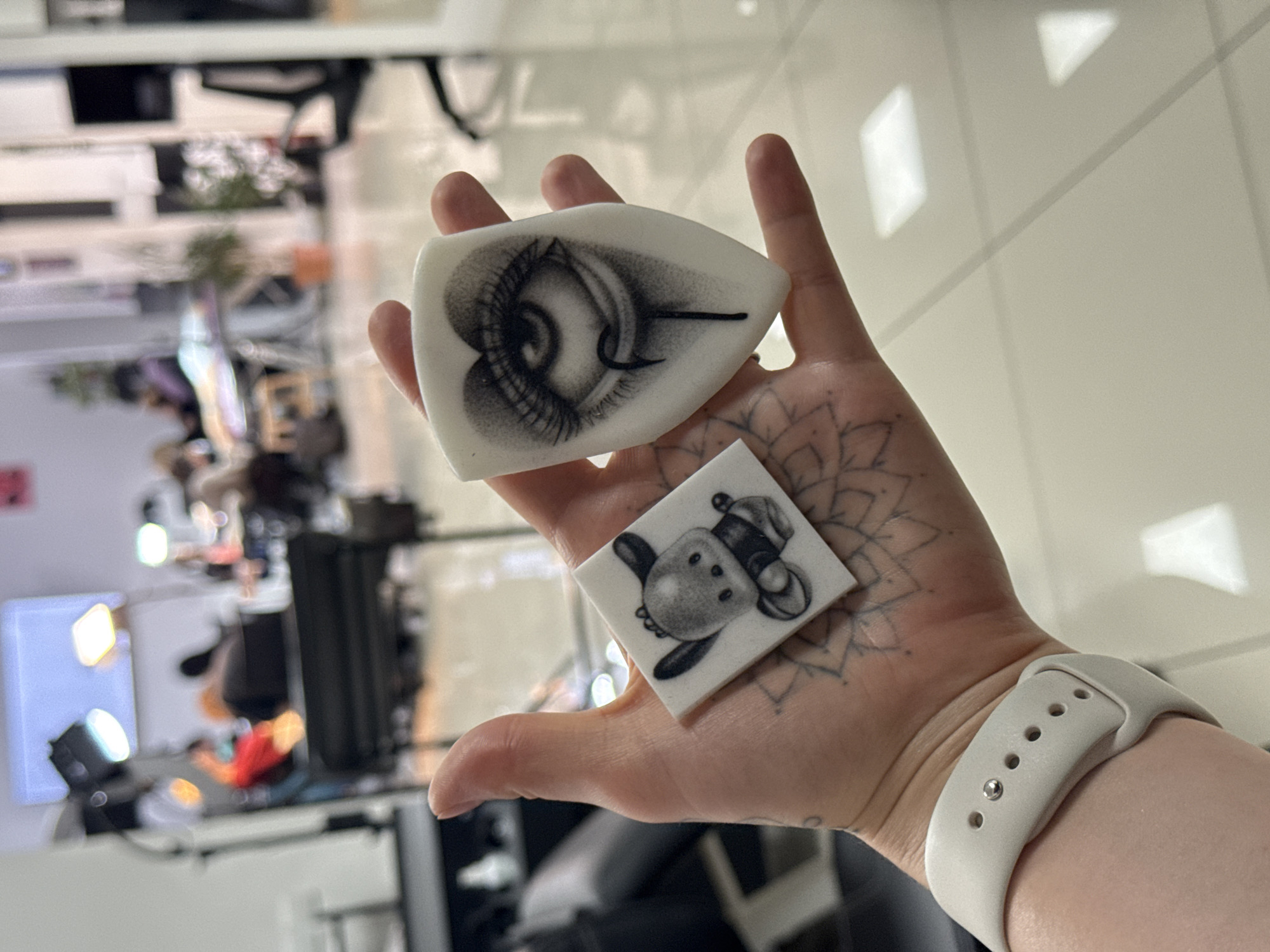 A hand holds two small, white tattoo practice skins with drawings on them. The skin closest to the fingertips has a rendering of an eye with a fishhook through it. The skin closer to the wrist has a rendering of a sheep wearing a hat. There is a mandala tattoo visible on the hand. The wrist has a white Apple watch band on it. The background is blurry but appears to be a cluttered tattoo shop or studio. There is no evidence to suggest this image was taken in Rostov-on-Don, Russia.