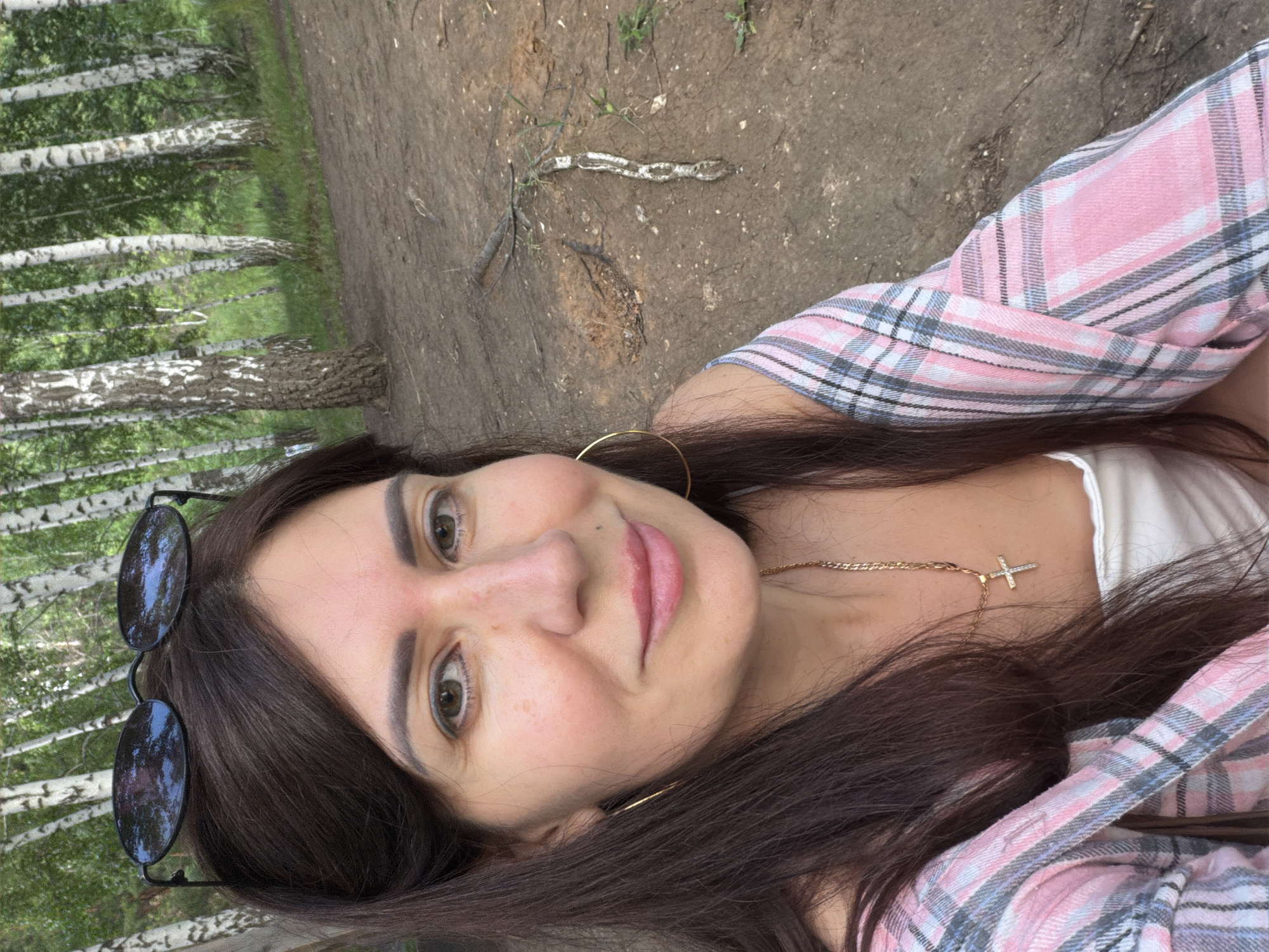 A woman is taking a selfie outdoors in a birch forest. She has long, dark hair and is wearing sunglasses on top of her head. She is also wearing a plaid shirt over a white top and a gold chain necklace with a cross pendant. The background is a dense forest of birch trees with a dirt ground. The image appears to be taken in the city of Volzhskiy, Russia, likely in a park or recreational area.