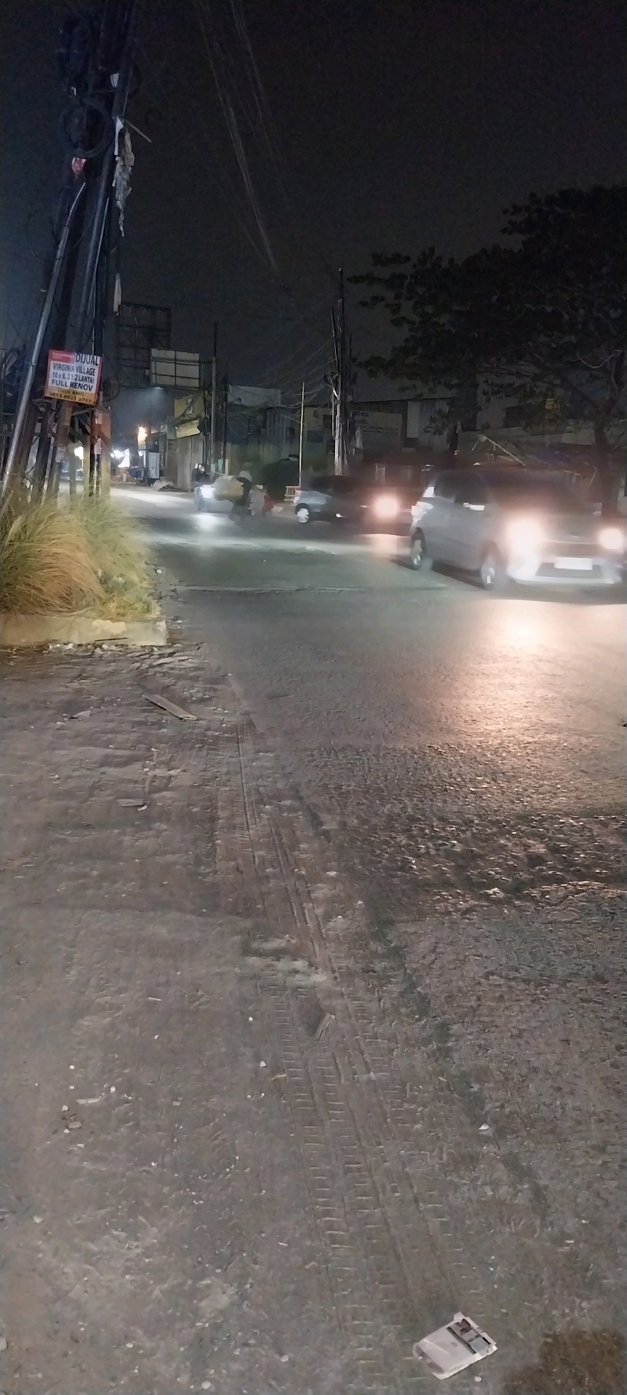 An exterior, nighttime, vertical, medium shot shows a street scene in Kelapa Dua, Indonesia. The foreground is a paved surface with tire tracks. In the middle ground, two cars are driving, one light-colored and one darker-colored. The light-colored car is in the right foreground, and its headlights are on. The darker-colored car is further back and to the left, also with its headlights on. There are several people visible walking or standing near the cars. In the background, there are buildings and trees. A sign on a pole on the left side of the image reads "DIJUAL VIRGINIA VILLAGE 10X21 2 LANTAI FULL RENOV."