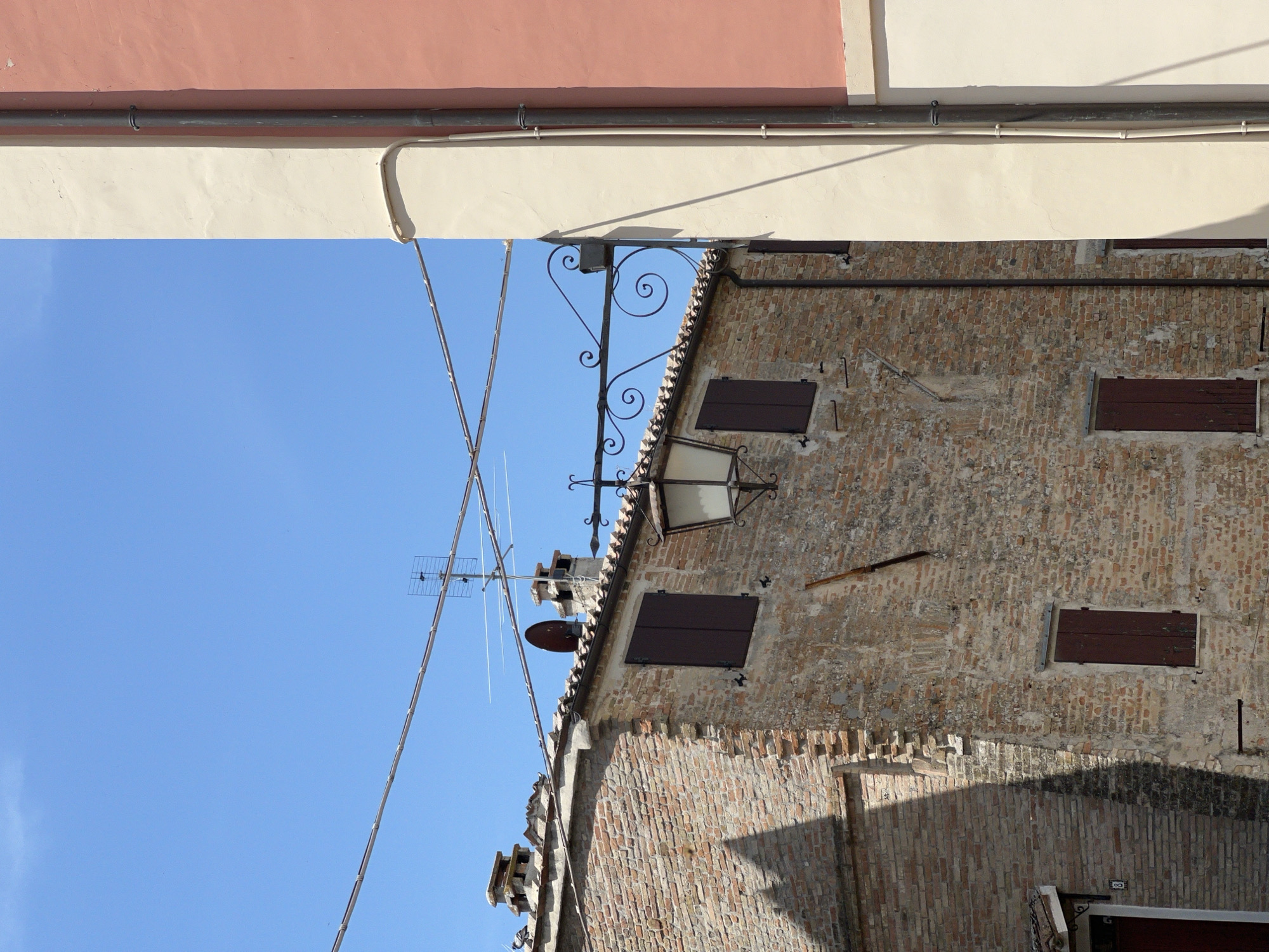 The image depicts an external view of an old brick building in Barbara, Italy. The building has a tiled roof and several dark brown shutters on its windows. A vintage-style lantern hangs from an ornate wrought-iron bracket attached to a pole. The building's brickwork shows signs of age and weathering.
In the foreground and right side of the image, a section of another building is visible, painted in two distinct colors: a light cream color and a pinkish-orange color. A dark downspout runs vertically along the cream-colored section, with several clips securing it to the wall.
The sky above is a clear, bright blue. Several wires crisscross the sky, and an antenna is mounted on the roof of the brick building. The overall scene suggests a sunny day in a historic Italian town.