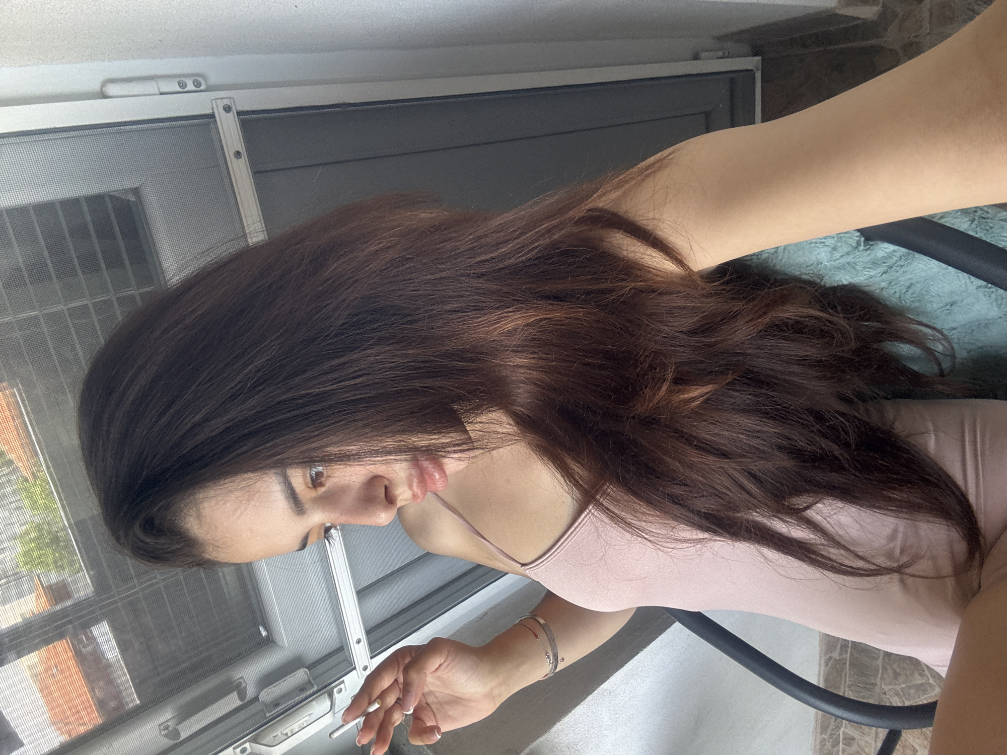 A young woman is depicted in a side profile, looking downwards, in what appears to be an indoor setting next to a window in Plovdiv, Bulgaria. Her long, dark brown hair, which has visible auburn or reddish-brown highlights, flows over her shoulders. She is wearing a light pink top, possibly a tank top or bodysuit.
Her left hand is visible, holding a lit cigarette between her fingers. Two thin, metallic bracelets are worn on her left wrist, and her fingernails have a neat manicure with white tips. The background reveals a window with a metal frame and a fine mesh screen. Through the screen, an outdoor scene is visible, including parts of a building with a reddish-brown, brick-like facade and green foliage from trees, suggesting a clear, bright daylight.
A black chair back is partially visible to her right, and a textured, possibly green, fabric object is beneath her arm. The flooring appears to be light-colored tiles. The woman's expression is neutral and focused downwards as she holds the cigarette. There is no visible text in the image.