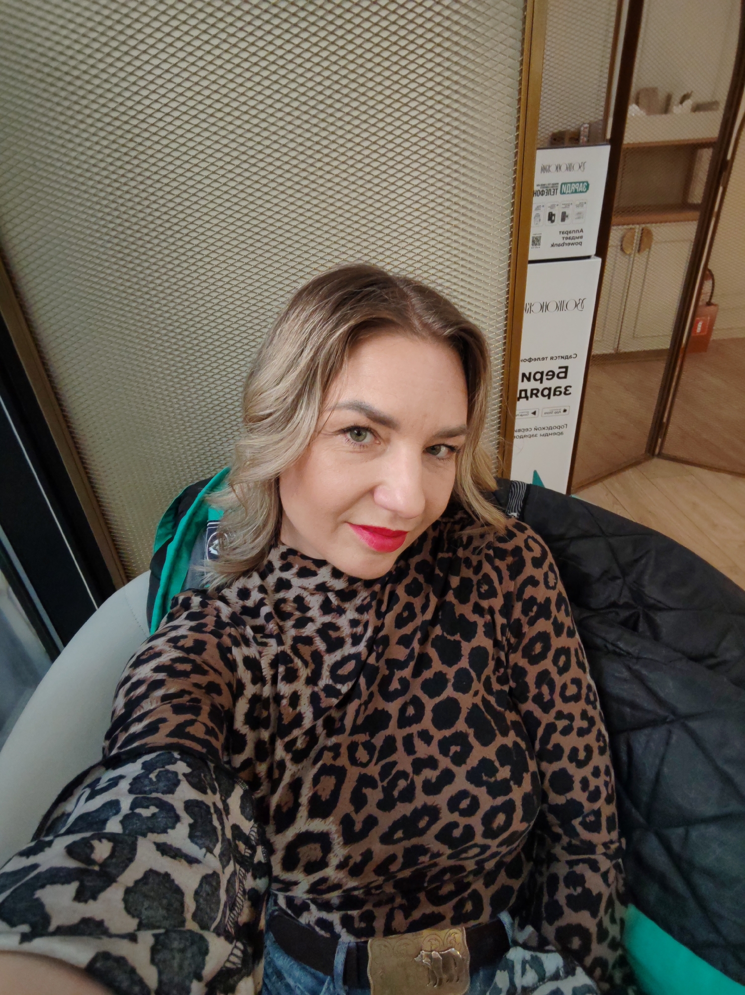 An indoor, eye-level selfie shows a woman with shoulder-length blonde hair and red lipstick, looking directly at the camera. She wears a long-sleeved, high-necked top with a brown and black leopard print pattern. Her lower body is partially visible, showing blue jeans and a belt with a prominent gold-colored buckle featuring a bear design.
She is seated in a light-colored armchair or seat, with a black quilted jacket draped over another chair or part of her seat to her right. The background features a textured wall, possibly a mesh or patterned wallpaper, in a light beige or off-white color. To her right, a wooden frame runs vertically, behind which is a mirrored surface reflecting parts of a room, including what appears to be shelves, a white cabinet, and a red object (possibly a fire extinguisher or bag).
Several signs are visible on the mirrored surface, displaying Cyrillic text and symbols, including what looks like QR codes. Due to the reflection, the text is reversed and largely indecipherable, though some words may relate to services or information, given the icons. The overall setting suggests a public or commercial indoor space, such as a cafe, waiting area, or office lobby, located in Saint Petersburg, Russia. The woman appears calm with a slight smile.