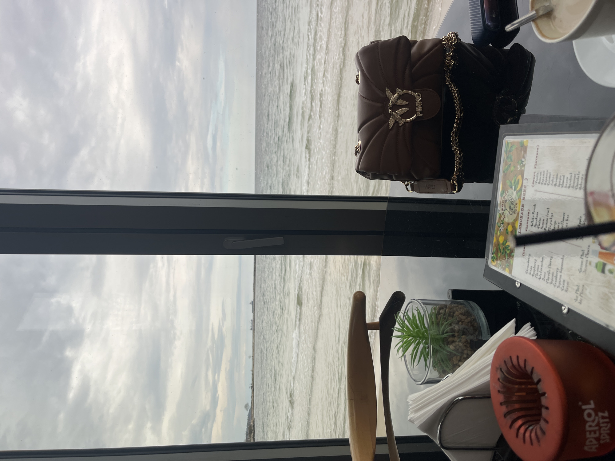 The scene is an interior view from a building overlooking the sea in Varna, Bulgaria, as the menu on the table contains text in Bulgarian. The overcast sky is visible through the window. On the table, there is a brown leather handbag with a gold chain and a Pinko logo, a black bag, a cup of coffee, and a menu card. Next to the menu, there is a napkin holder with an "Aperol Spritz" logo, a small decorative plant in a glass container, and part of a chair. The scene suggests someone is sitting at a table near a window with a view of the sea.