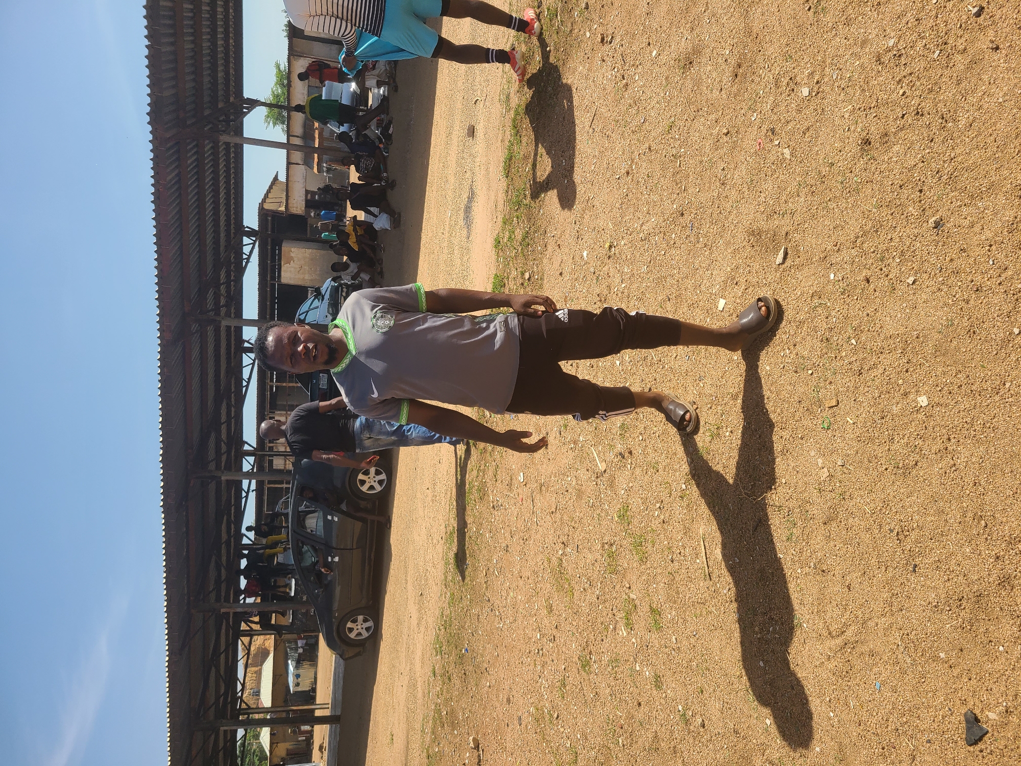 A man in a gray t-shirt and brown shorts stands on a gravel lot. He is looking towards the camera. He is wearing brown sandals. There are several cars parked in the background under a metal structure. There are other people in the background sitting and standing near the cars. The sky is blue. The location is Unknown, Unknown.