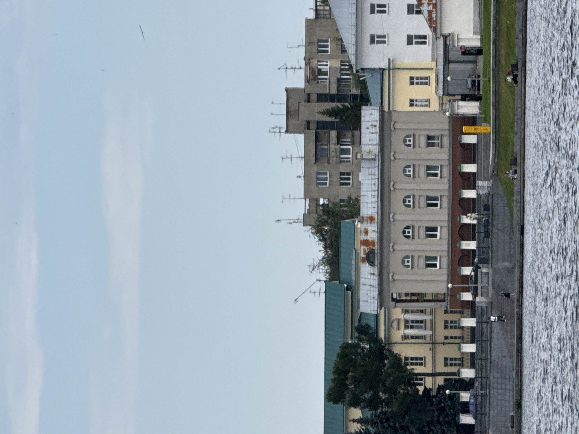 The image depicts a waterfront view of buildings and a body of water, likely a river or canal. The architecture suggests an urban environment in Yekaterinburg, Russia, given the style of the buildings, including a mix of older classical-style structures with grand facades and more utilitarian, modern apartment blocks with numerous antennas. The sky is overcast with light clouds.
In the foreground, a rippling body of water occupies the bottom portion of the image. A stone embankment or quay runs along the edge of the water. On the embankment, several individuals are visible. Two people are walking along the embankment, one dressed in a white shirt and dark pants, the other in a dark dress. Further along the quay, a few other individuals are seated on what appear to be benches.
Behind the quay, a row of buildings is situated. The closest building to the water has a light-colored facade with columns and arched window details, topped by a dark green roof. Adjacent to this are other buildings, including a pale yellow structure and a larger, grey building with multiple floors and numerous windows, many adorned with antennas. A white building with dark-framed windows is also visible on the right side. Some of the buildings appear to have undergone renovations or have aging elements, such as the rusty sections on one roof.
Vegetation is present in the form of trees, some dark green and coniferous, positioned behind the buildings. The overall impression is a city scene with a significant waterway, typical of a European city, and a blend of architectural periods. The weather appears to be temperate, with the overcast sky suggesting it could be a cool or mild day.