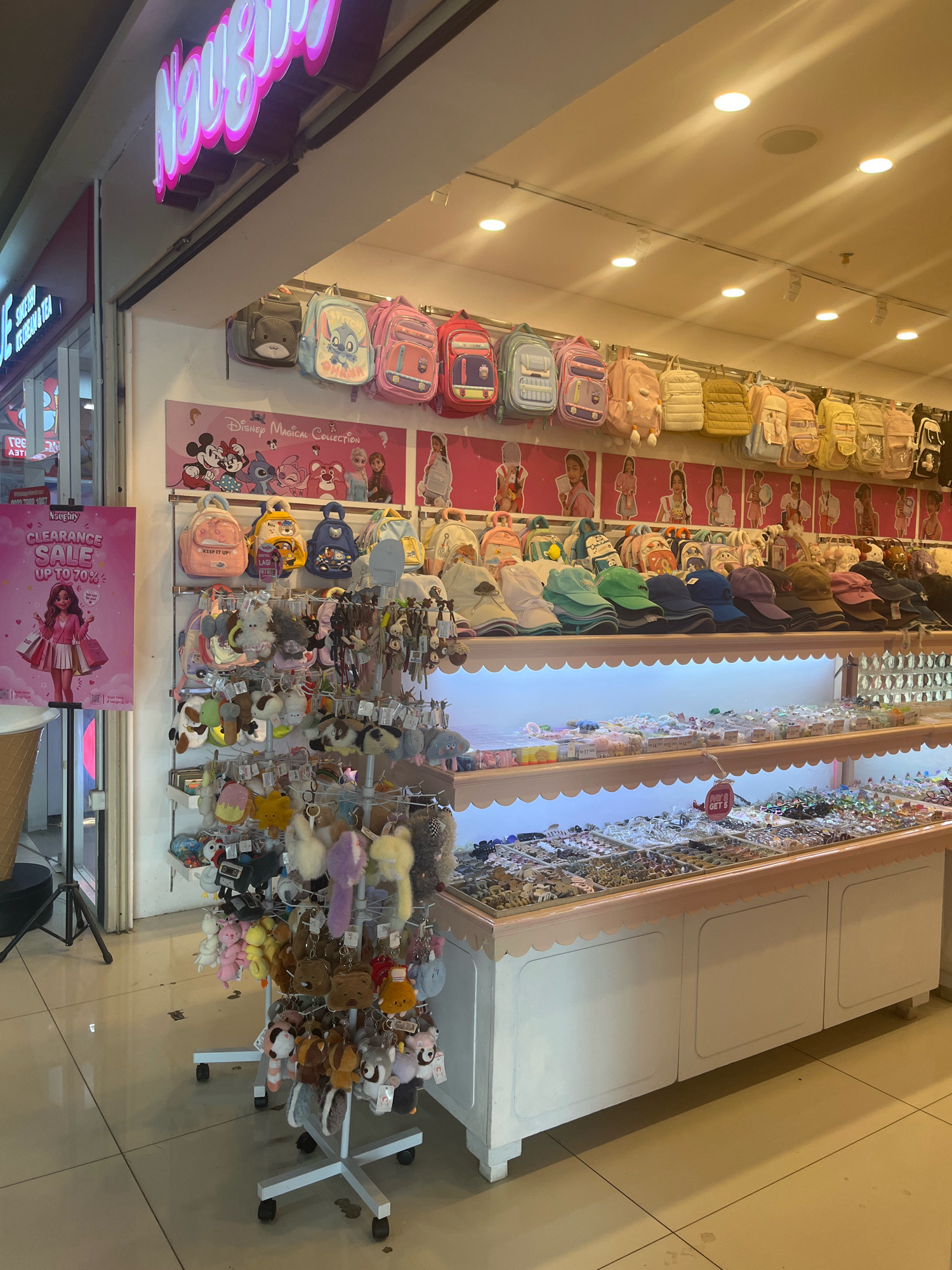 A brightly lit store interior, likely a boutique or specialty shop, features an abundance of merchandise organized on shelves and racks. In the foreground, a rolling display rack is laden with an assortment of colorful plush keychains and small stuffed animals. Behind this rack, shelves are filled with various accessories, including a large selection of baseball caps and hair accessories. Higher shelves display an array of backpacks, many of which feature cartoon characters, suggesting a focus on children's or novelty items. A prominent pink sign with illuminated white lettering reading "Naughty" is visible at the top of the image, and a pink promotional poster advertising a "Clearance Sale UP TO 70%" is affixed to a stand on the left. The store appears clean and well-maintained, with tiled flooring. The overall atmosphere is cheerful and vibrant, with the merchandise suggesting a target demographic of young people or those interested in cute and whimsical items. The location is identified as Sengkang, Indonesia.