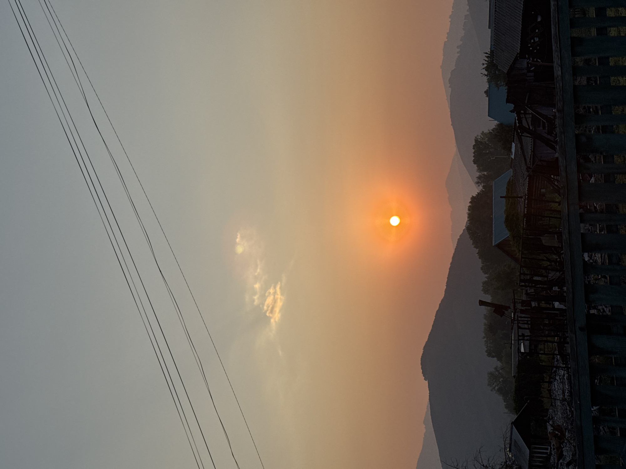 This image captures a serene sunset scene in the village of Soloneshnoye, Russia. The sun, a radiant orb of orange, is setting behind a range of hazy, layered mountains, casting a warm glow over the sky. A few wispy clouds are scattered across the horizon, catching the sunlight and adding depth to the scene. In the foreground, silhouetted against the vibrant sky, are the rooftops of houses and structures, hinting at a rural settlement. Power lines stretch across the upper portion of the image, a subtle reminder of civilization amidst the natural beauty. The overall atmosphere is tranquil and picturesque, evoking a sense of peace and solitude.