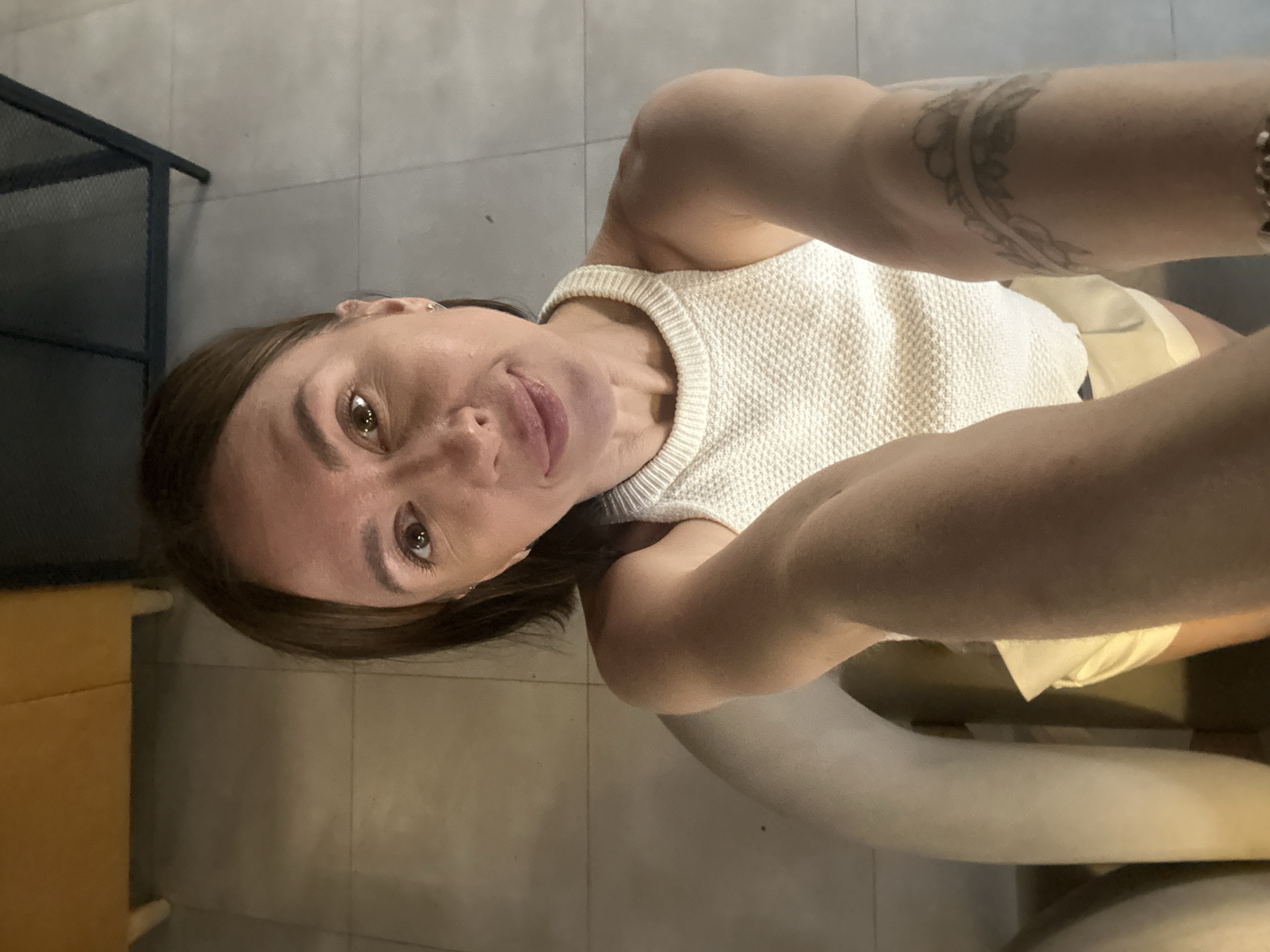 An overhead, close-up selfie shows a woman with light brown hair and fair skin. She is wearing a white, sleeveless, knit top with a high neckline and light-colored shorts. Her left arm is extended towards the camera, and her right arm is visible at an angle. A tattoo is visible on her right arm. She is sitting on a light-colored chair in what appears to be an indoor setting with tiled flooring. The background includes a portion of a yellow piece of furniture and a dark metal structure. There are no identifiable landmarks or contextual clues that would place this image in Votkinsk, Russia.