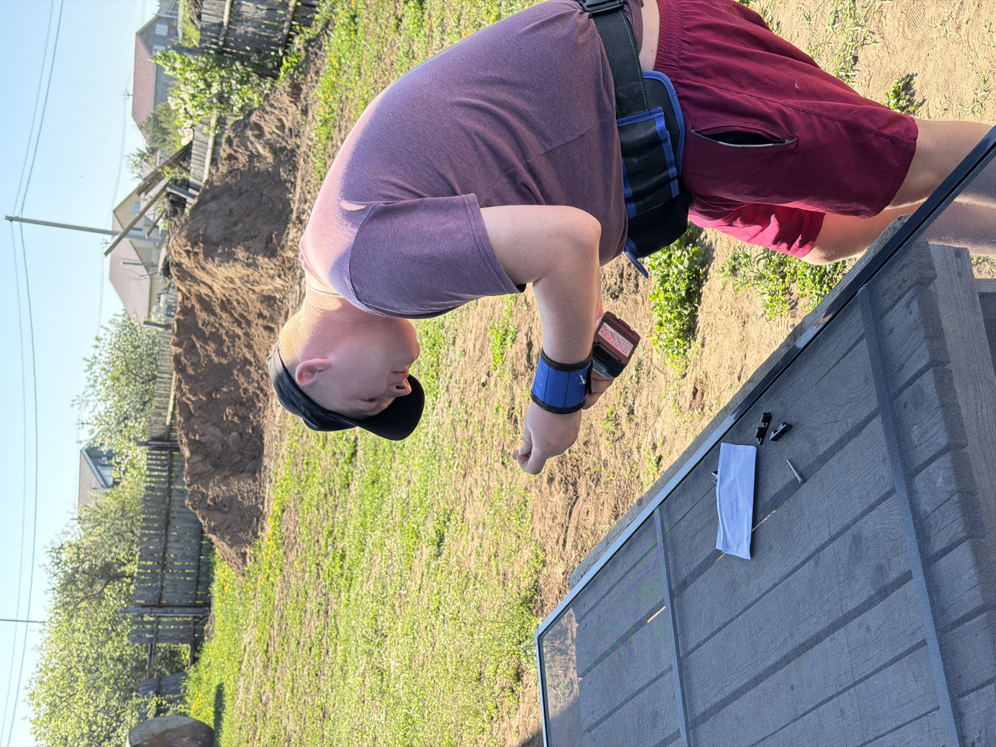 A fair-skinned person with a shaved head, wearing a purple t-shirt, burgundy shorts, a black baseball cap, and a weightlifting belt, is standing in a grassy yard. They are bending over and looking at items on a gray wooden surface, which includes a small white packet, a nail, and a few small black objects. The person is wearing a blue wrist support. In the background, there is a wooden fence, a pile of dirt, some trees, and several houses. The location is Chaikovsky, Russia.