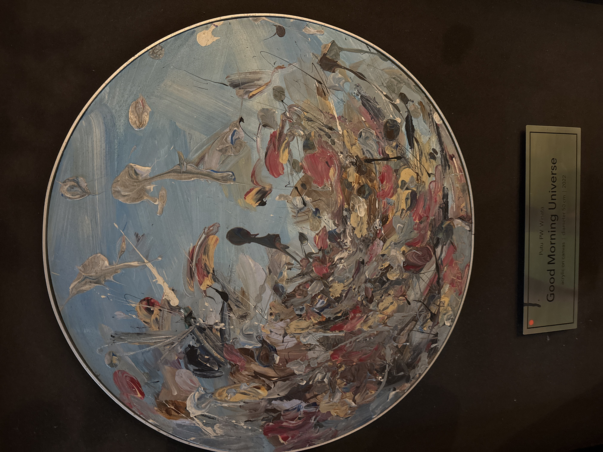 This media file features a circular abstract painting with a predominantly light blue background, suggesting an open sky or perhaps a celestial scene. The painting is rendered in thick, expressive brushstrokes of various colors including white, gray, brown, red, and yellow. The abstract forms suggest movement and energy, possibly depicting a cosmic event or the dynamism of nature. On the right side of the image, a small, rectangular plaque is visible, providing details about the artwork. The text on the plaque reads: "Putu PW Winata", "Good Morning Universe", and "acrylic on canvas | diameter 50 cm | 2022". The painting itself is presented against a dark, possibly velvet, background, which serves to highlight the vibrant colors and textures of the artwork. The setting appears to be an indoor exhibition or gallery space, given the display method of the artwork and the presence of the informational plaque. The image captures the artwork from a slightly angled perspective, with the circular canvas taking center stage.
