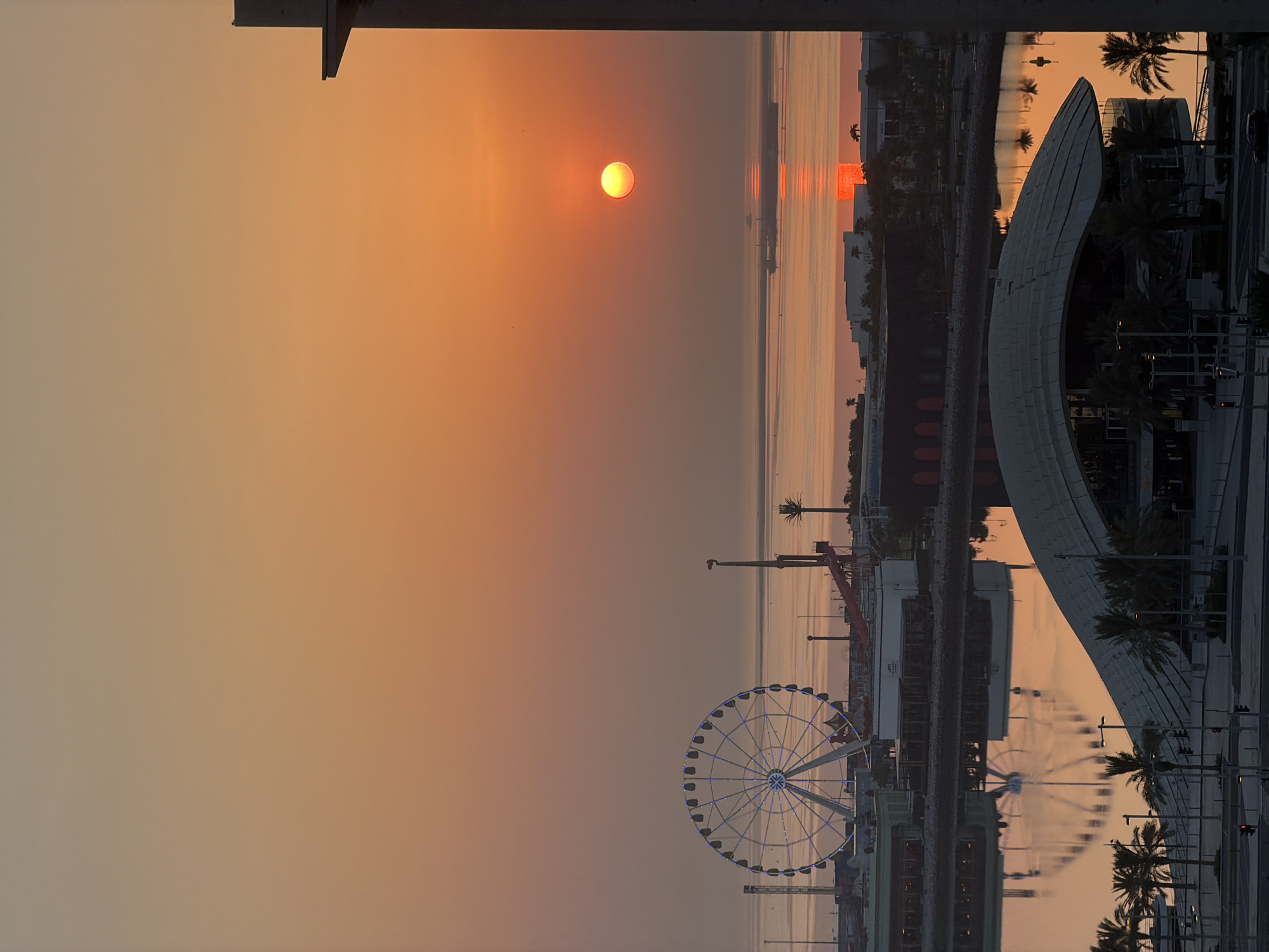The image depicts a sunset over the city of Lusail, Qatar. The sun, a bright orange orb, is low on the horizon, casting a warm glow across the sky and the water. In the foreground, a modern building with a curved roof is visible, surrounded by palm trees. A body of water reflects the sunset and the surrounding structures. In the mid-ground, a Ferris wheel with blue lights illuminates a section of the waterfront. Beyond that, a calm sea stretches to the horizon, with a few ships visible in the distance. The overall scene is serene and bathed in the golden hues of twilight.