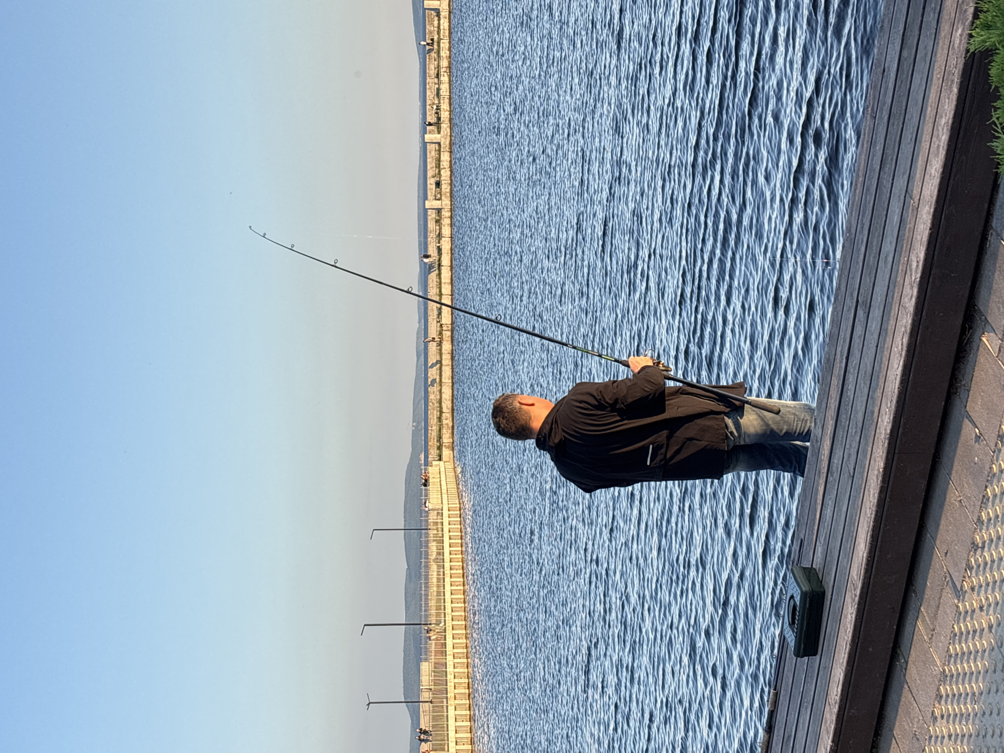This media file depicts a man fishing on a wooden pier overlooking a body of water. The man, wearing a black jacket and blue jeans, is holding a fishing rod. A small green tackle box sits on the pier next to him. The water is calm with gentle ripples. In the background, a long promenade with lampposts stretches along the coastline, with hills or mountains visible in the distance. The sky is clear and blue, suggesting a pleasant day. The lighting indicates it is likely late afternoon or early evening. The scene is peaceful and suggests a recreational activity. While the specific location is not explicitly visible, the provided context places it in Tolyatti, Russia. There is no visible text in the image.