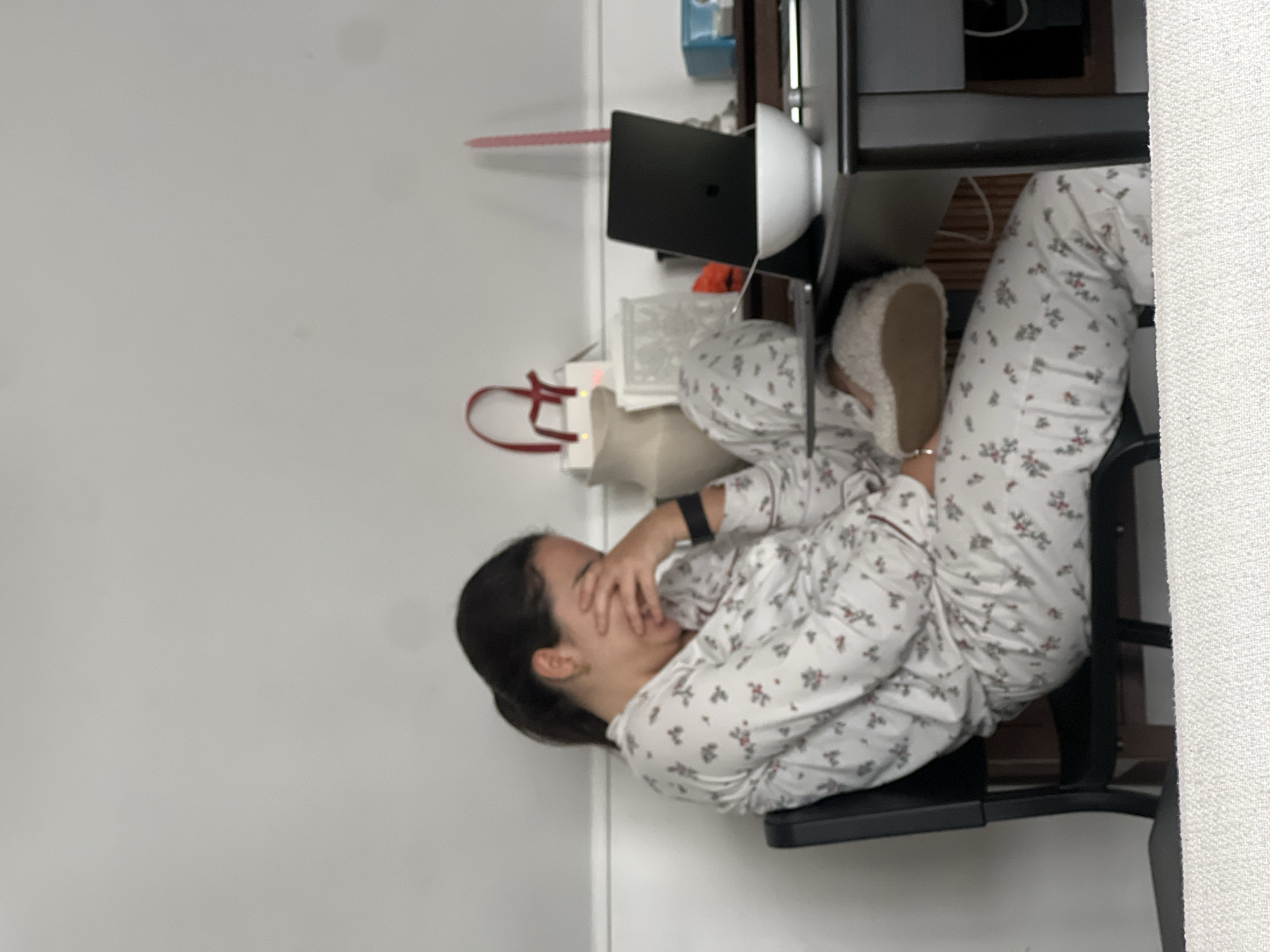 A woman in floral pajamas sits at a desk in what appears to be an indoor room with white walls. Her face is partially obscured by her hands. A laptop, a white bowl, and other objects are visible on the desk. No information suggests the location is Paris, France.