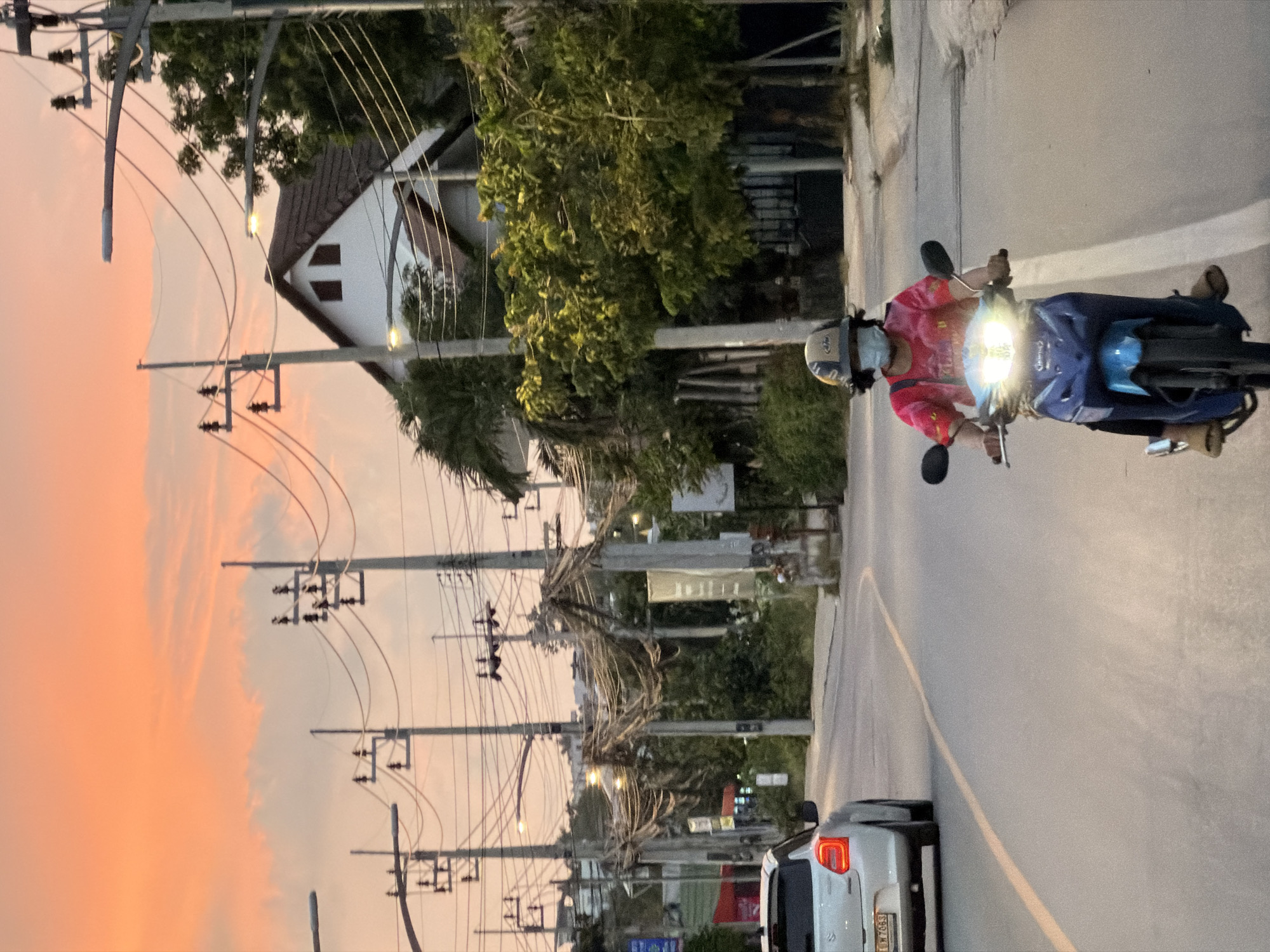 A person wearing a pink and yellow tie-dye shirt, a blue helmet, and a light blue face mask is riding a blue motorcycle on a paved road in Ko Samui District, Thailand. The motorcycle's headlight is on, illuminating the road ahead. The person's feet are visible, wearing light brown sandals. Behind the motorcycle, a white car is driving away from the camera, its red taillights illuminated. The road has a yellow line marking the lane. The environment is urban, with numerous utility poles and electrical wires lining the street. A white house with a dark-tiled roof and two small windows is visible in the background, surrounded by green trees and foliage. The sky is a soft orange hue, indicative of a sunset.