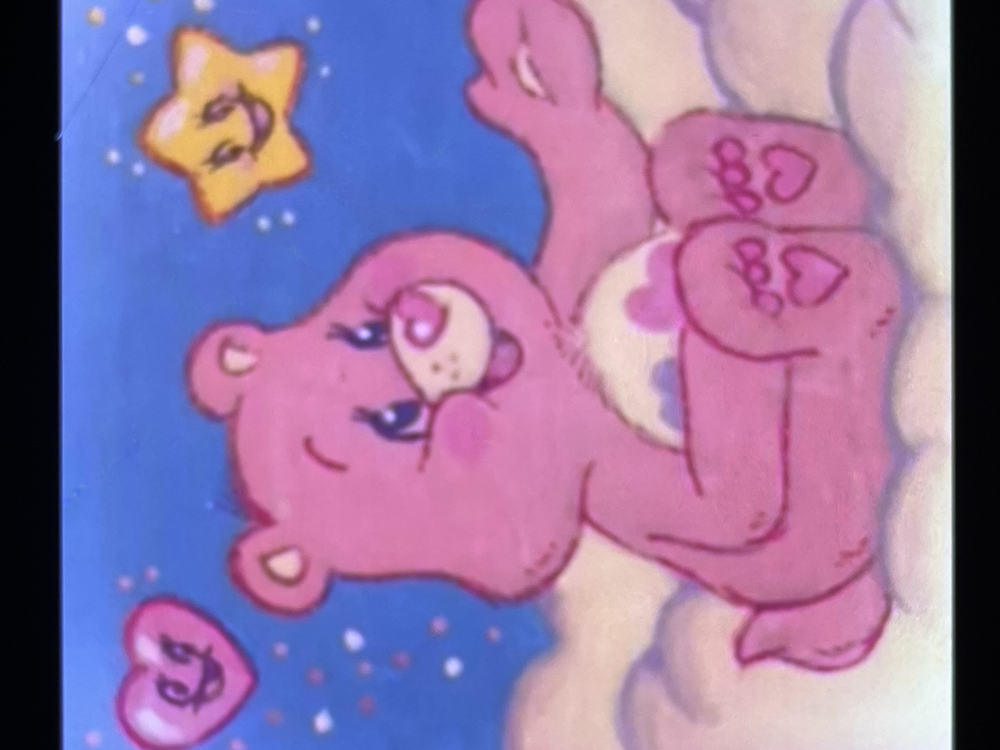 The image shows a stylized pink bear floating amongst clouds and stars. The bear has a cheerful expression with rosy cheeks. Above the bear, a yellow star with a smiley face and rosy cheeks also appears to be floating. Below the bear, a pink heart with a smiley face is visible. The bear's belly has a light-colored symbol, possibly representing a heart shape with smaller hearts inside. The overall scene is whimsical and suggests a dreamy or magical setting, possibly a night sky given the presence of stars. The lighting suggests daytime or a bright, illuminated scene. The style of the artwork is reminiscent of children's cartoons or illustrations. There is no visible text in the image. The location context of Cadek, Indonesia, does not appear to be directly relevant to the content of the image itself, which depicts a universally recognizable cartoon character and motifs.