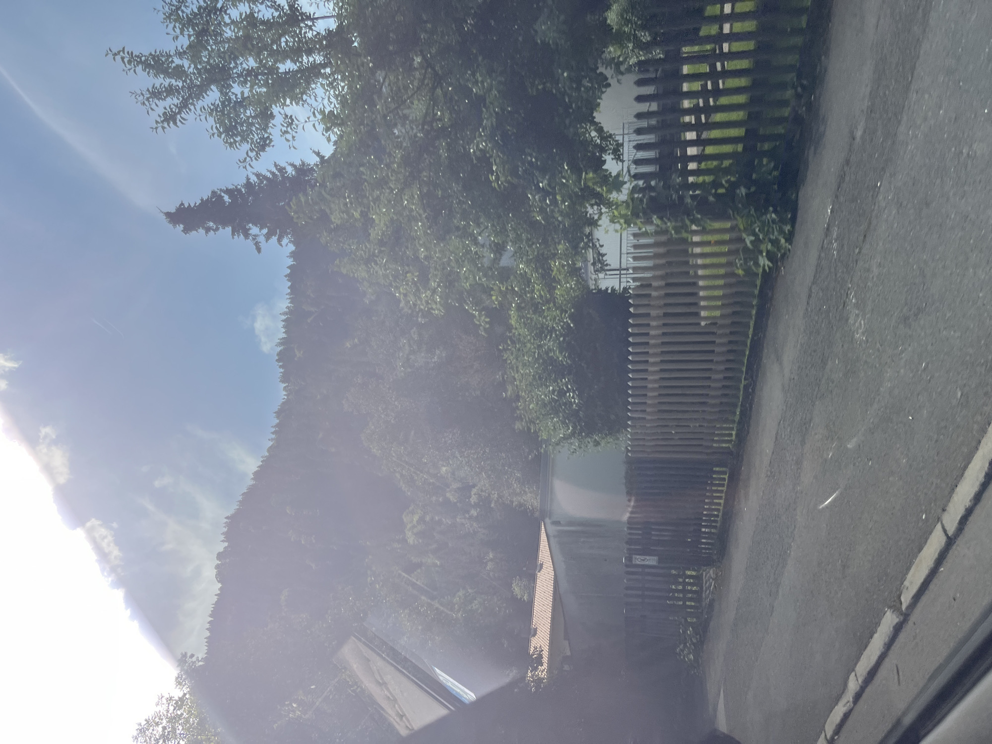 The image depicts a sun-drenched, slightly overexposed view of a residential area in Oberammergau, Germany. In the foreground, a paved road runs parallel to a wooden picket fence. Beyond the fence, a white building with a red-tiled roof is visible, partially obscured by trees. Behind the buildings, a densely wooded hillside rises, topped by a clear blue sky with some wispy clouds and sun flare. The overall impression is of a rural or suburban setting bathed in bright sunlight.