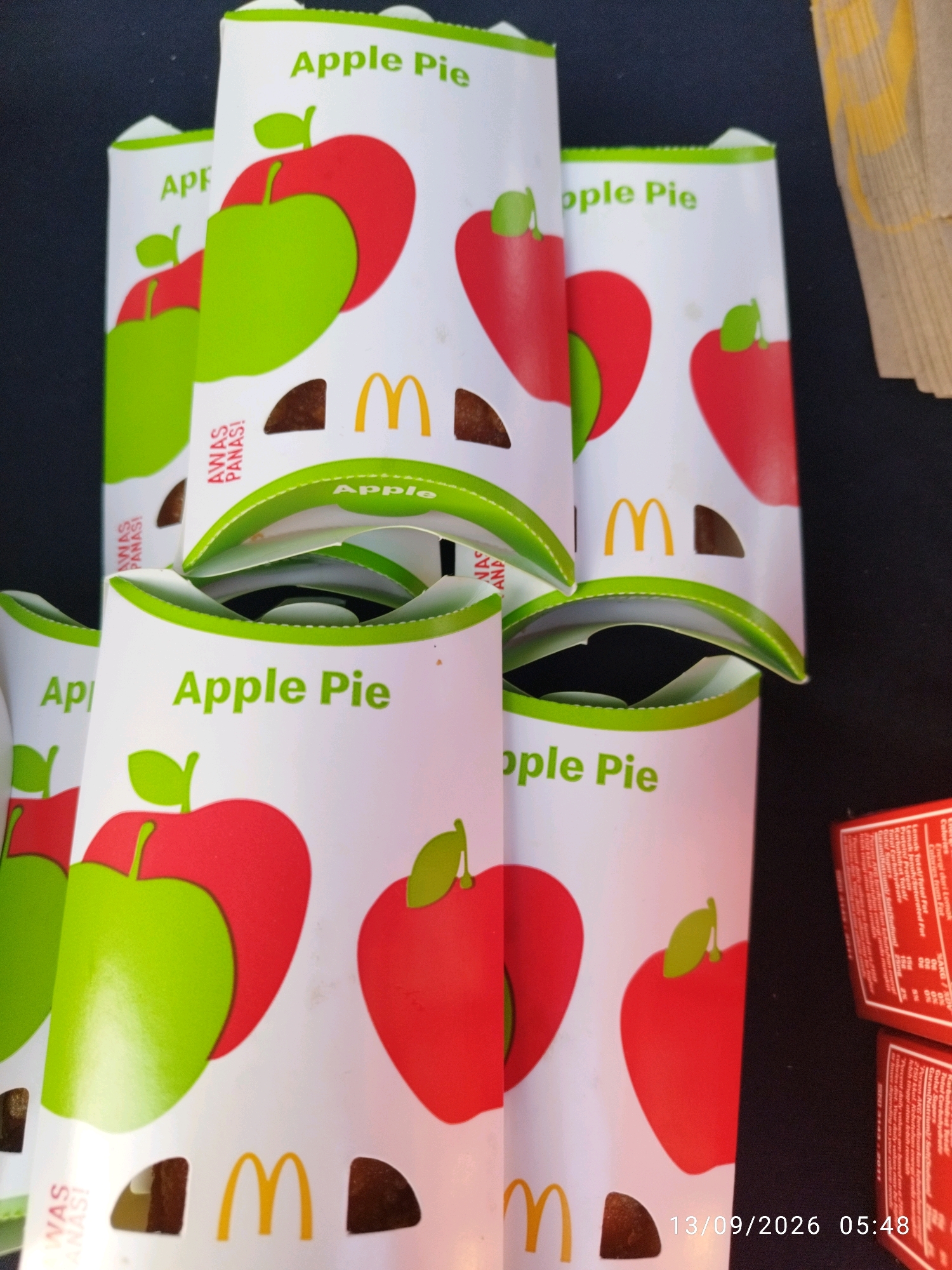 The image shows a stack of McDonald's apple pie boxes. The boxes are white with green and red apple illustrations. The "Apple Pie" text is in green. Some boxes have the McDonald's logo in yellow. The boxes are arranged vertically, with some of them open, revealing the apple pies inside. The background is dark. A date and time stamp at the bottom right corner reads "13/09/2026 05:48". The context of the image is believed to be Mojokerto, Indonesia, though this is not directly visible in the image itself.