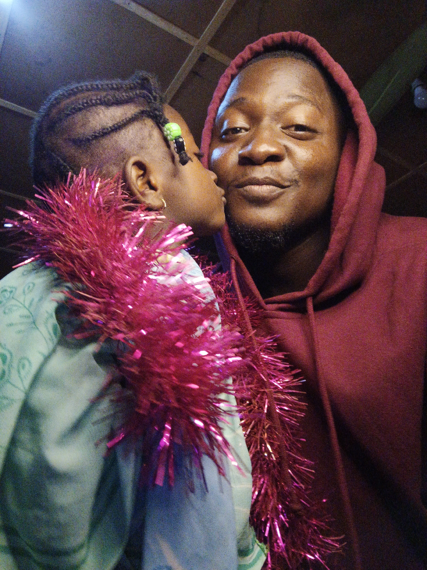 An adult male wearing a maroon hooded sweatshirt and a young person with braided hair are embracing and posing for a selfie. The young person is kissing the adult male's cheek. A bright pink, tinsel-like garland is draped around the young person's neck and obscures part of the adult's face and chest. The background is dark and appears to be an indoor setting with a wooden ceiling. The image is slightly grainy.