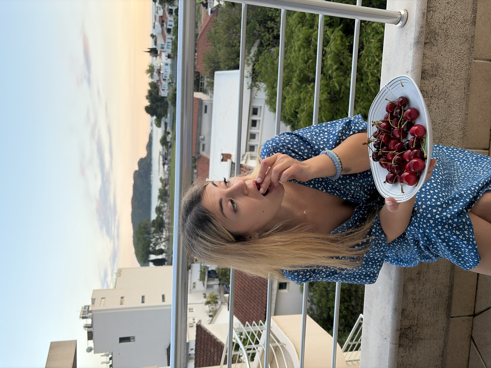 A fair-skinned woman with blonde hair is eating cherries from a white plate on a balcony. She is wearing a short-sleeved blue dress with white hearts. In the background is a view of Budva, Montenegro, with white buildings, trees, and the sea. The sky is a mix of blue, orange, and grey, indicating either sunrise or sunset. The balcony has a silver railing.