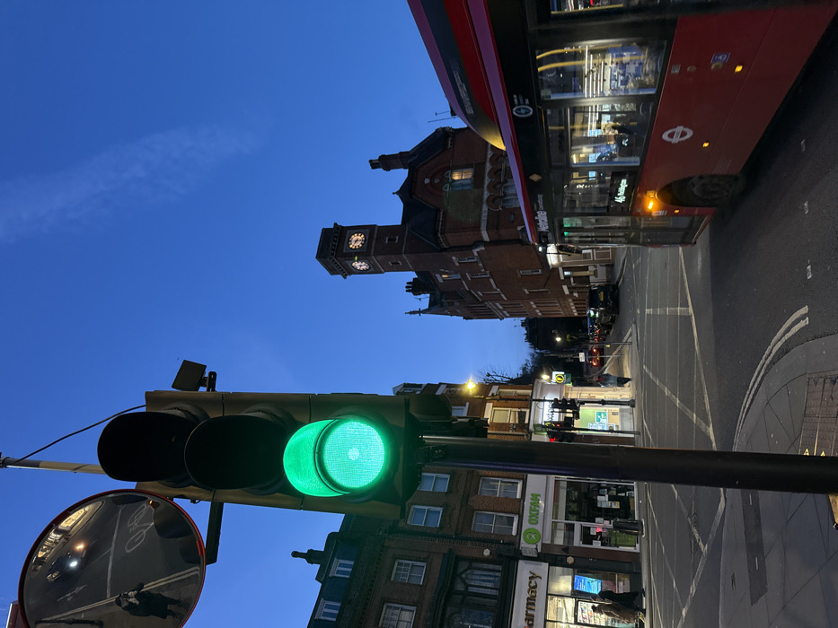 The image captures an urban street scene at dusk or early night, characterized by a deep blue sky and artificial illumination. The photograph appears to be taken from a low angle, looking upwards.
In the foreground, a prominent black traffic light is visible on the left side, with its bottom green signal brightly illuminated. The other two lights are off. Attached to the same pole as the traffic light, a circular convex mirror reflects a distorted view of the street and what appears to be a person holding a camera or phone, likely the photographer.
To the right and further back, a red double-decker bus, characteristic of London, is partially visible. The side of the bus shows reflections of interior lights and passengers. Text on the bus indicates "NatWest" and the route number "41". The bus appears to be turning or stopped at a junction.
In the mid-ground, a distinctive brick building with a clock tower stands against the blue sky. The architecture suggests an older, possibly Victorian, style with intricate details and multiple windows. The clock faces on the tower are visible, showing a time that appears to be approximately ten minutes past an hour.
Below the clock tower building and extending towards the bottom right of the image are other urban structures, featuring commercial establishments at ground level. Clearly visible signs include "Oxfam" with its green logo and "Pharmacy." The street surface, composed of asphalt, is visible in the lower right quadrant, displaying white road markings including cross-hatching and outlines of what might be a pedestrian crossing. Faint streetlights can be seen further down the road, enhancing the evening atmosphere. The combination of the red double-decker bus, specific architectural styles, and shop names suggests the location is likely in London, UK.