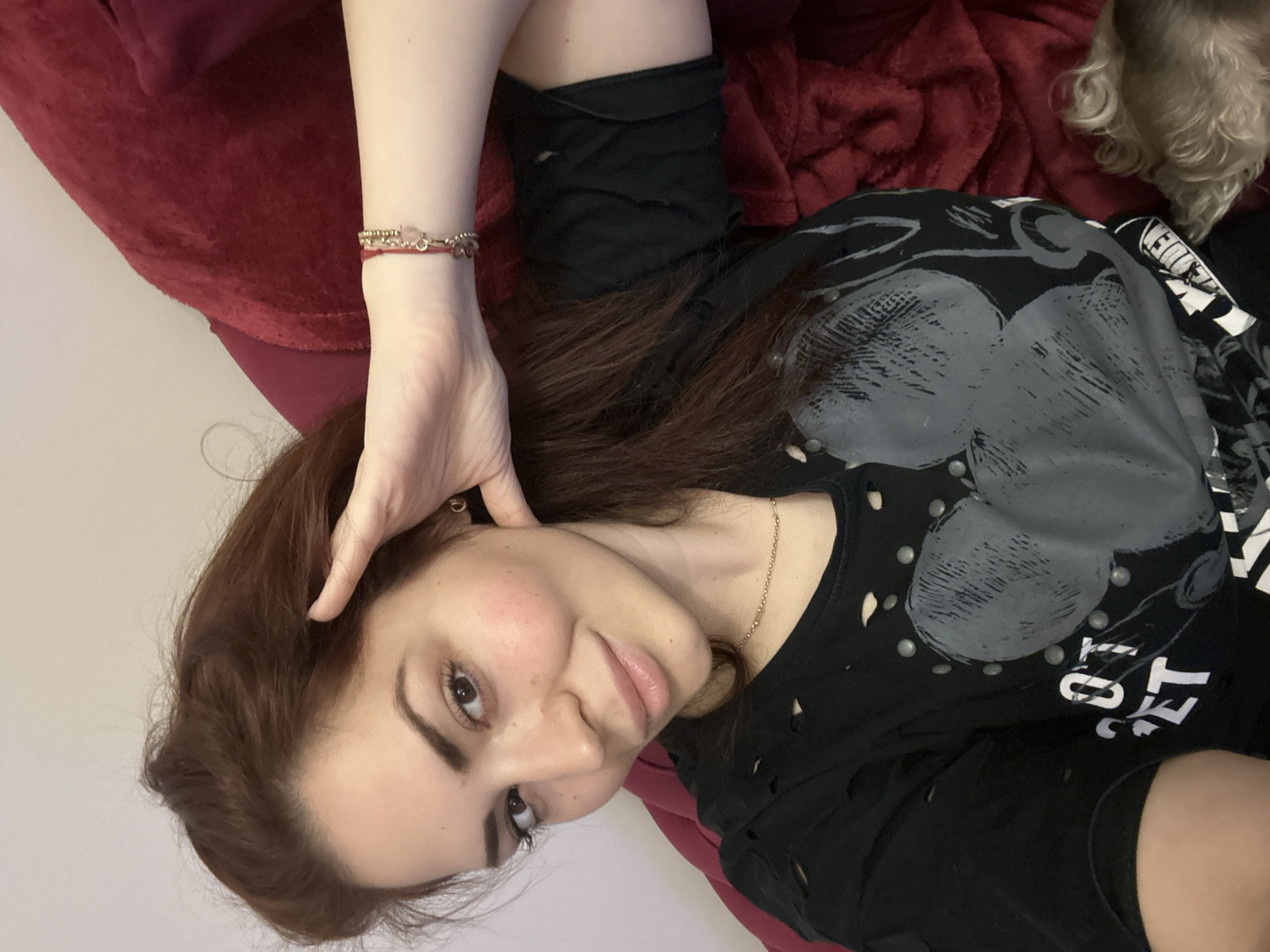 A young woman with long brown hair is lying down on a red surface, possibly a bed or couch. She is wearing a black shirt with a gray print and possible stud detailing. She has a bracelet on her left wrist and a necklace around her neck. Her left hand is near her head. A small dog is partially visible in the upper right corner of the image. The background is a plain light color, possibly a wall. The location of the photograph cannot be determined to be in Sofia, Bulgaria.