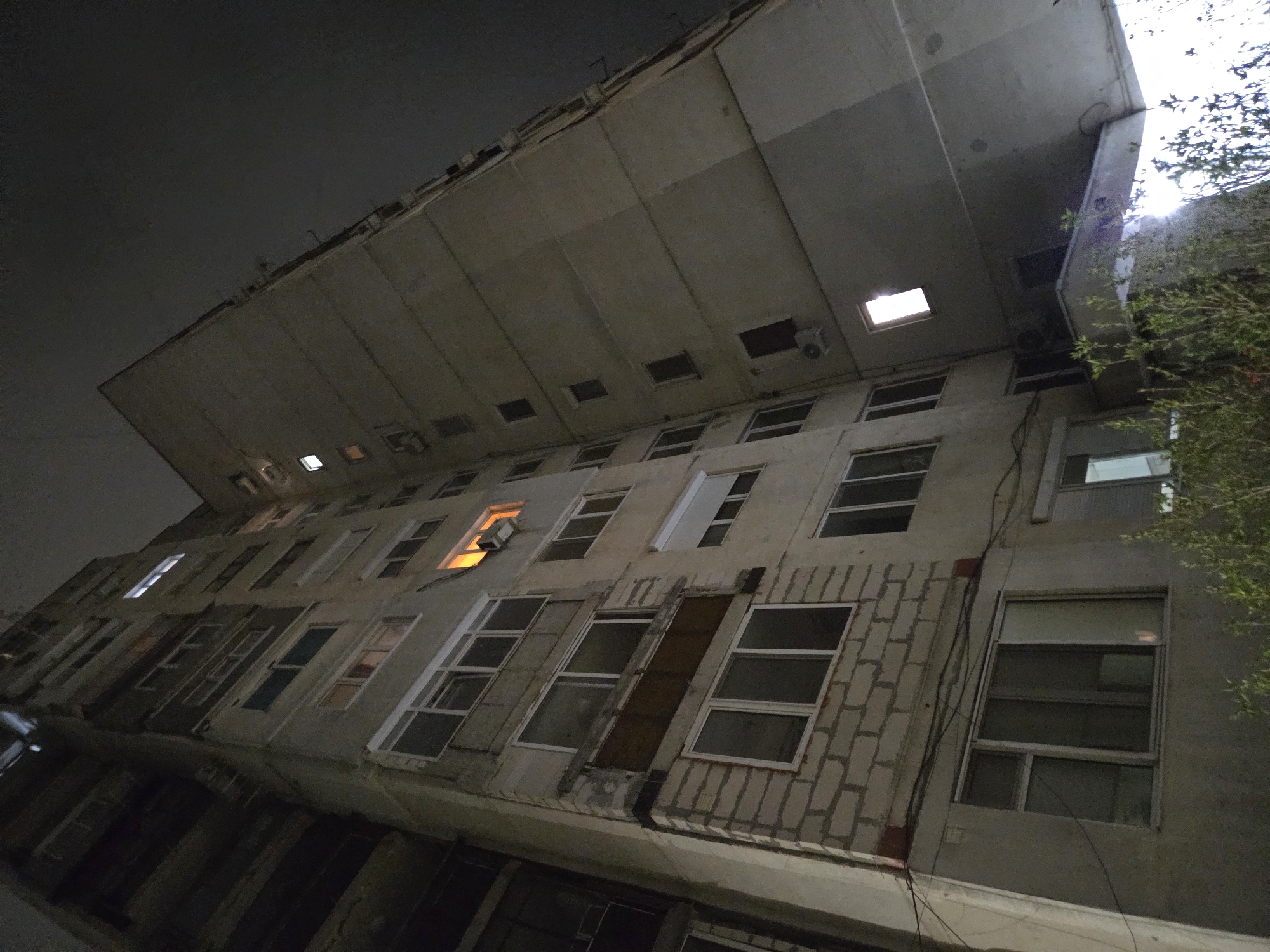 The image depicts a low-angle view of a concrete building at night. The building appears multi-storied with rows of windows, some illuminated from within, and some blocked off or boarded up. The exterior shows signs of wear and possible disrepair, with patching apparent on the facade. Electrical wires run along the building's exterior. The architecture includes an overhanging ledge, likely for balconies. To the right of the image, foliage from a tree or bush obscures part of the building and sky. The sky is dark, with a faint light source near the top right corner, possibly from streetlights or the moon. The location is not specified in the image, and no people, subjects, or interactions are visible. The building's environment suggests an urban setting.