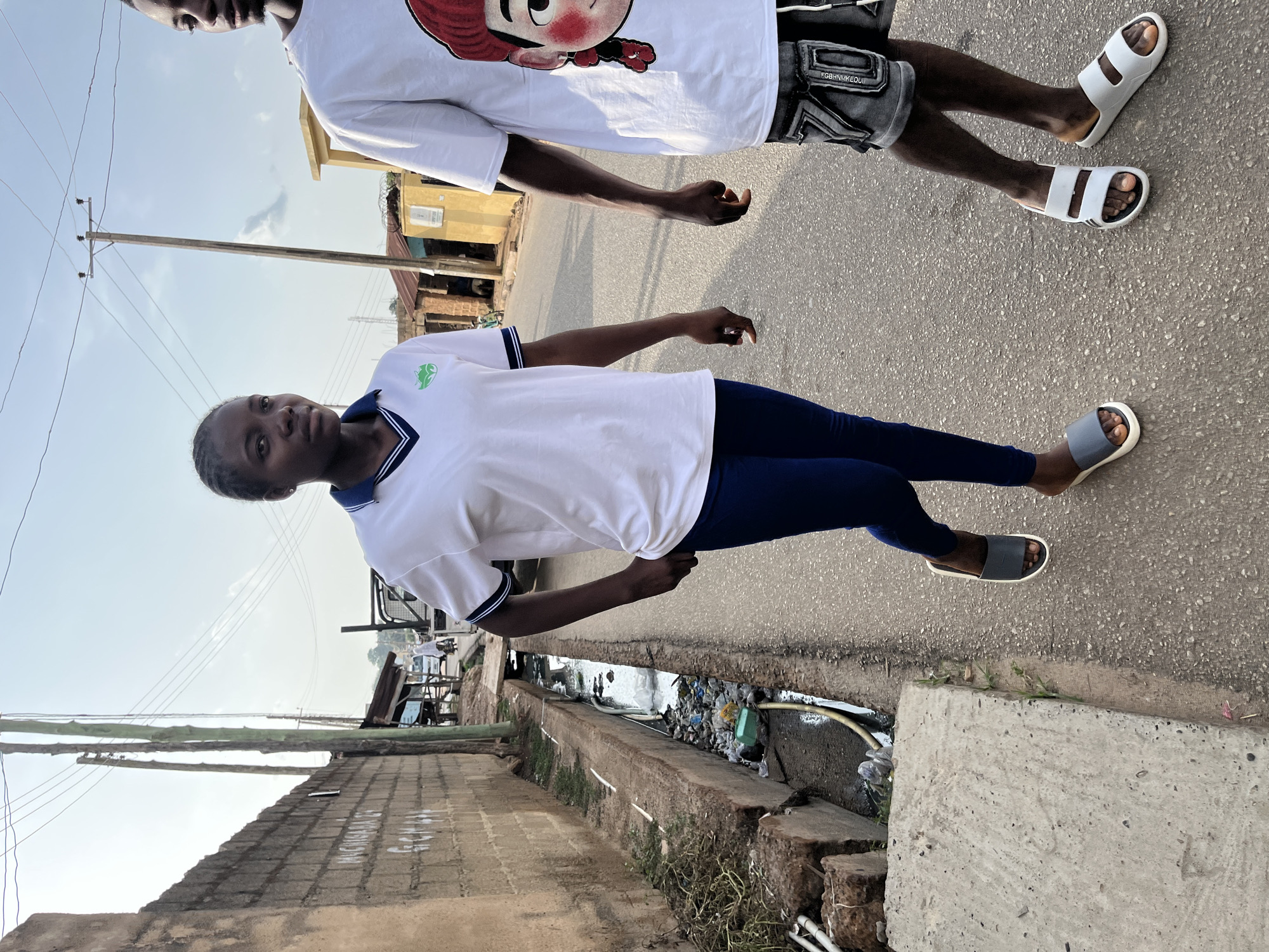 The image shows two people standing on a paved street in Jos, Nigeria. The first person is wearing a white t-shirt with a cartoon design and grey shorts. The second person is wearing a white polo shirt with blue trim and blue leggings. Both are wearing sandals. A concrete drainage ditch runs alongside the street, filled with trash and debris. Buildings are visible in the background, along with utility poles and wires. The sky is clear and the scene is lit by daylight.