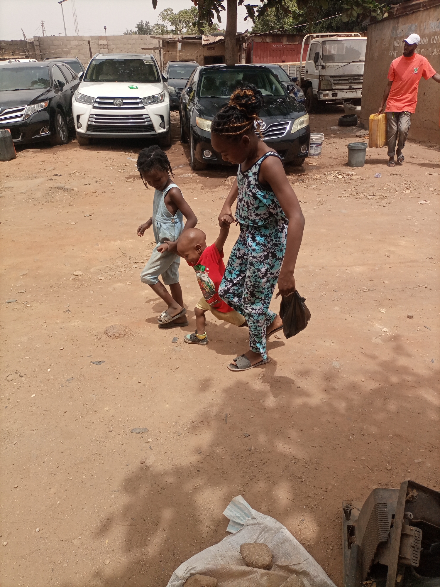 The image depicts a dirt-covered open area in Kaduna South, Nigeria. There are several vehicles, including two black SUVs, a white SUV, and a white truck in the background.
In the foreground, three children are walking hand-in-hand: a young girl in a floral jumpsuit holds hands with a younger boy in a red t-shirt, and a second young girl in denim overalls walks alongside them. A man in an orange shirt, white cap, and khaki pants walks in the background, carrying a yellow jerrycan. A bucket is on the ground near him.
The ground is predominantly dirt with scattered debris. In the lower right corner, there is a partially visible bag filled with something, and what appears to be a damaged piece of machinery or appliance.