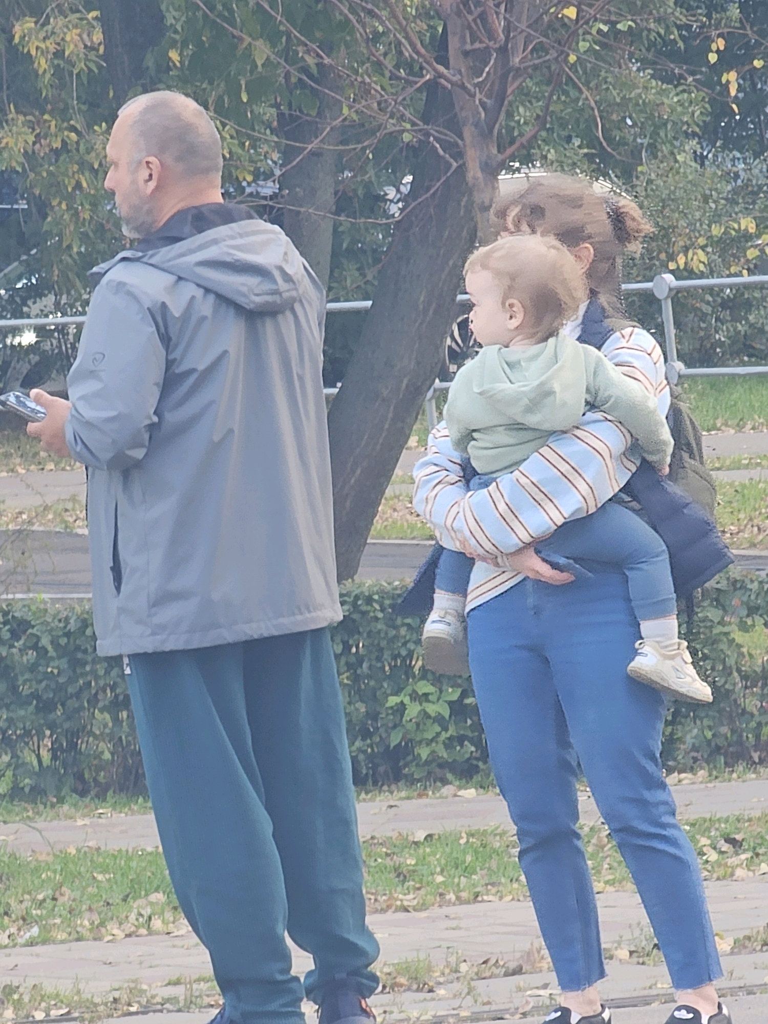 In a city park in Khimki, Russia, a bald man with a beard stands to the left, holding a mobile phone. He wears a grey hooded jacket and teal sweatpants. To his right, a woman holds a young child. The woman is partially obscured, but wears a striped long-sleeved shirt, blue pants, and a dark vest. The child is dressed in a light green hooded sweatshirt. The background shows trees and foliage, suggesting an outdoor, possibly autumnal, setting.