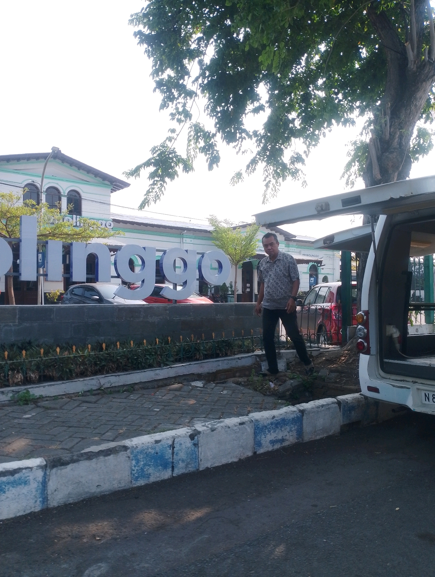 A medium shot captures a man standing in front of a sign that reads "PROBOLINGGG". The man is wearing a short-sleeved patterned shirt and dark pants. Behind him, a white vehicle with its rear door open is parked. The sign stands on a low stone wall with plants in front of it. In the background, a building with arched windows and a white and green facade is visible, along with trees and other vehicles. The foreground shows a sidewalk with blue and white painted curbs. The scene appears to be an outdoor public space in the city of Probolinggo, Indonesia.