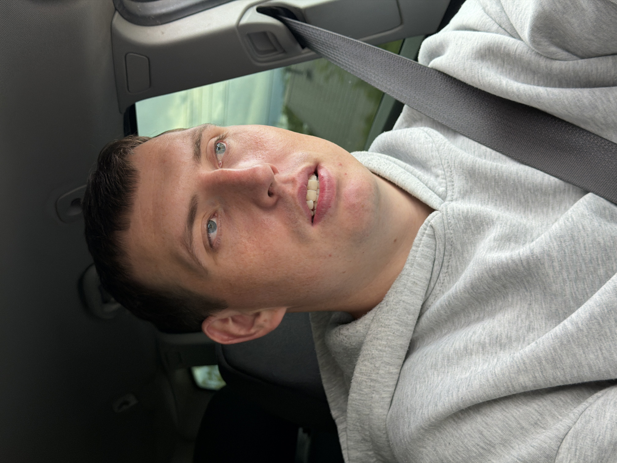 A close-up, slightly rotated portrait of a young man with short dark hair and blue eyes. He is wearing a gray hooded sweatshirt and is secured with a seatbelt. The interior of a car is visible in the background, with a blurred view of buildings and greenery outside the window. The exact location within Chaikovsky, Russia cannot be determined from the given image.
