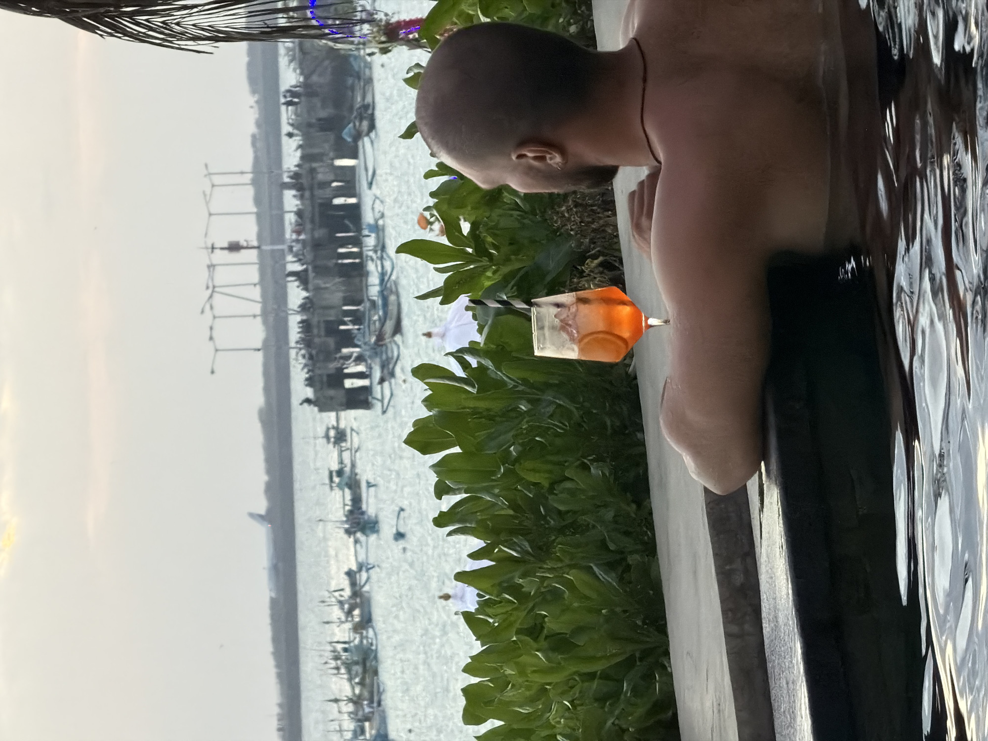 A man with a shaved head, wearing a necklace, is relaxing in a pool of water. He is facing away from the camera, looking out at a body of water. In the water, there are numerous small fishing boats and a large pier with lights and people. In the distance, an airplane is visible in the sky. In the foreground, a glass of orange-colored liquid with ice and orange slices sits on the edge of the pool, next to a black and white striped straw. Lush green foliage is visible behind the pool. The scene appears to be set in Kuta, Indonesia.