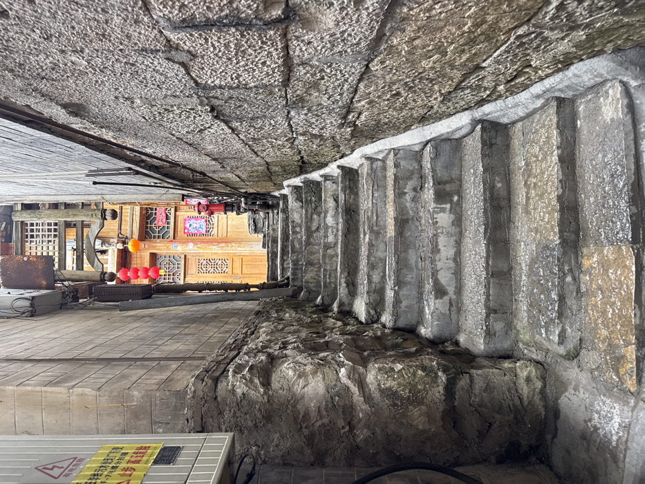 This image captures a steep, narrow stone and concrete stairwell in Furong, China, ascending between two tall, aged buildings. The foreground features a worn, damp concrete staircase with approximately ten visible steps leading upwards. The left wall consists of a natural, moss-covered rock formation at the base, transitioning to a grey block wall above, which has a grey utility box with a yellow warning sign in Chinese characters affixed to it. The right wall is constructed from rough-hewn, irregular stone blocks. At the top of the stairs, a traditional Chinese wooden building with intricately carved double doors is visible, featuring latticework above. Overhead, a cluster of three red round lanterns and one yellow spherical lantern hang. To the right of the doors, a red fire hydrant is mounted on a small landing. No individuals are present in the scene.