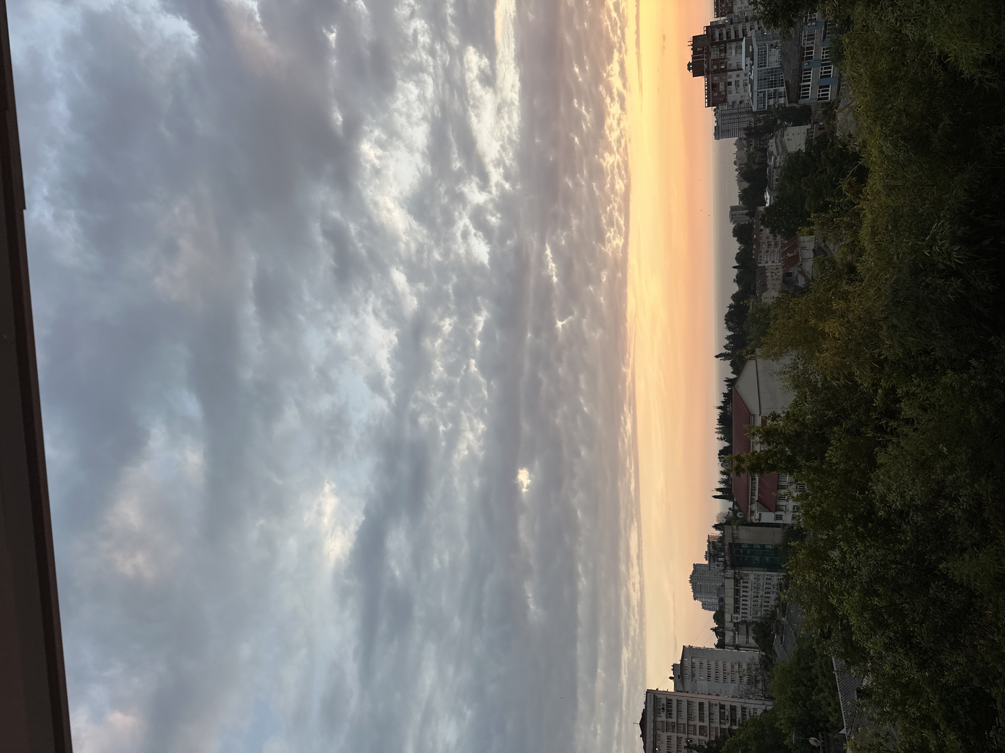 The image shows a cityscape during a sunset, likely in Sochi, Russia, given the architectural styles of the buildings. The sky is filled with clouds, ranging from light gray at the top to hues of orange and yellow near the horizon, indicating the setting sun. Below the sky, a dense layer of dark green trees forms the foreground, obscuring much of the lower portion of the city.
Several buildings are visible, ranging in height and architectural style. On the left side, there is a tall, multi-story apartment building with many windows. To its right, other apartment buildings and residential structures are clustered together, some appearing older with balconies and others more modern. In the mid-ground and background, more buildings are visible, interspersed with trees. The buildings appear to be located on hills or slopes, characteristic of Sochi.
In the distance, beyond the buildings, a body of water is visible, likely the Black Sea, with a faint hazy horizon line where the sky meets the water. The overall impression is a view of a coastal city at dusk, with a mix of urban development and abundant greenery. No people or specific activities are depicted in the image. The scene is static and captures a moment in time.