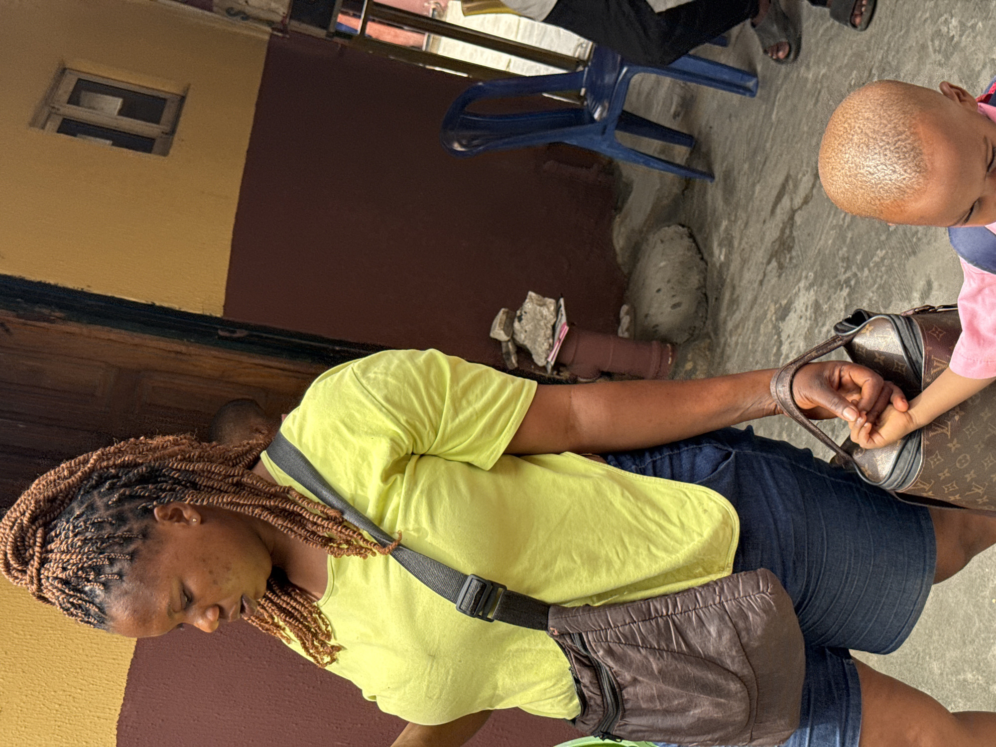 A woman with braided brown hair wearing a yellow t-shirt and blue denim shorts is holding the hand of a child. The child, wearing pink clothing, holds a Louis Vuitton handbag. The woman has a brown fanny pack strapped across her chest. They stand on a gray concrete surface. Behind them is a brown and yellow wall with a small window. In the background, an adult sits on a blue plastic chair. The scene appears to be set in an urban environment, likely Ado, Nigeria.