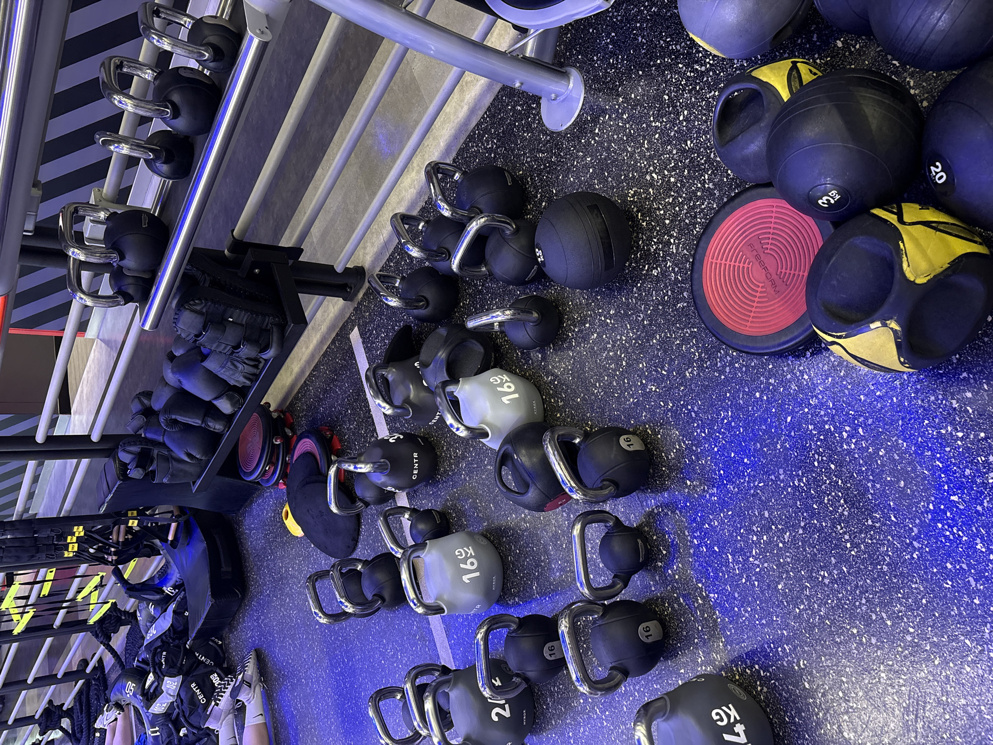 This image shows a collection of gym equipment laid out on a speckled blue rubber floor, with blue lighting casting a shadow. The scene is likely indoors, in a fitness facility. The equipment includes various kettlebells and medicine balls, some with visible weight markings like "16 KG" and "20". There are also some boxing gloves and other training gear stored on a rack. In the lower part of the image, a person's legs and feet wearing athletic shoes are visible, suggesting someone is present or has recently been in the area. The lighting suggests it might be evening or a time with artificial lighting. There is also some visible text, including brand names like "CENTR" and "FreeFORM", and weight indicators. The setting appears to be a gym in Nong Pa Khrang, Thailand. The overall impression is one of a functional workout space.
