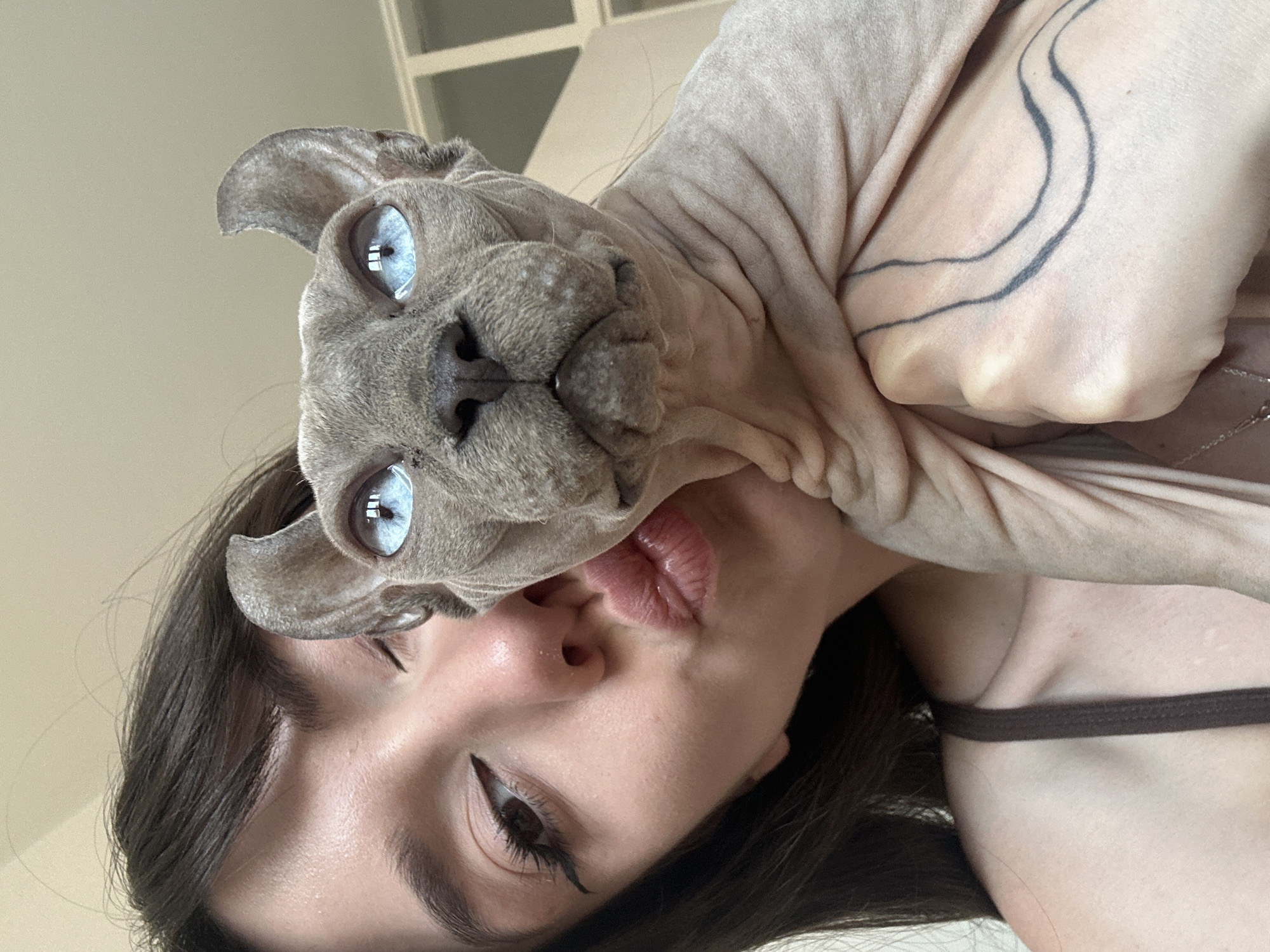 A close-up selfie shows a young woman with dark hair and winged eyeliner puckering her lips towards the camera. She is holding a hairless cat with striking blue eyes close to her face. The cat's skin is a light gray, and it has large, prominent ears. The woman is wearing a dark brown tank top, and a tattoo is visible on her arm, partially obscured by the cat. The background is out of focus, but appears to be an indoor setting with natural light. The location is not specified in the image.