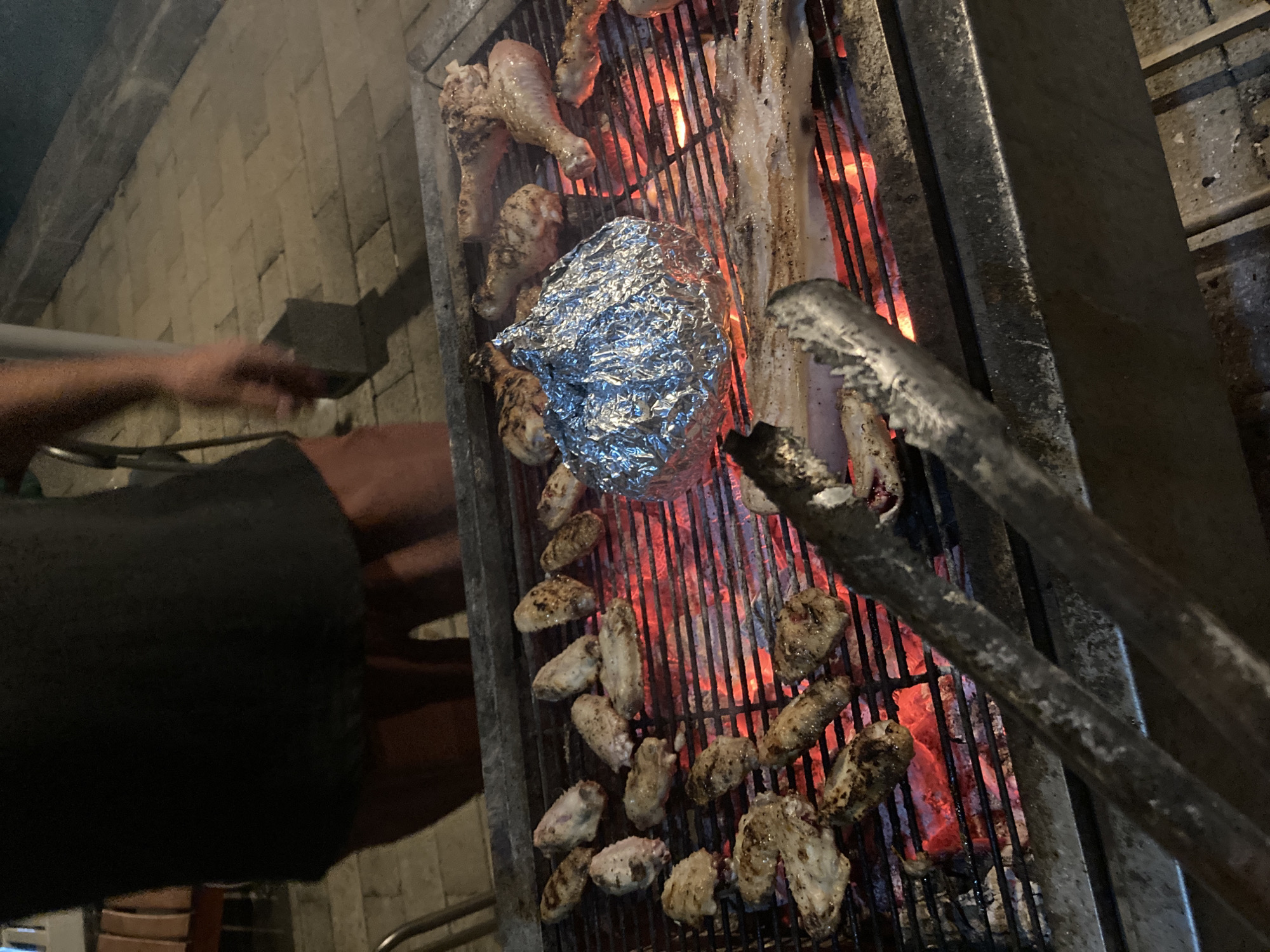 Captured at an outdoor BBQ location in Mũi Né, Vietnam, the image focuses on a grill loaded with food. A blurred human arm and hand appear on the left, near a person wearing a dark-colored shirt. A metal grill is crowded with chicken legs, chicken wings, unidentified white strips of food, and a foil-wrapped item. A set of metal tongs, heavily used and showing signs of wear, are positioned over the grill. Coals below the grill bars glow red, indicating high heat. The scene is bordered by what appears to be a brick or stone structure.